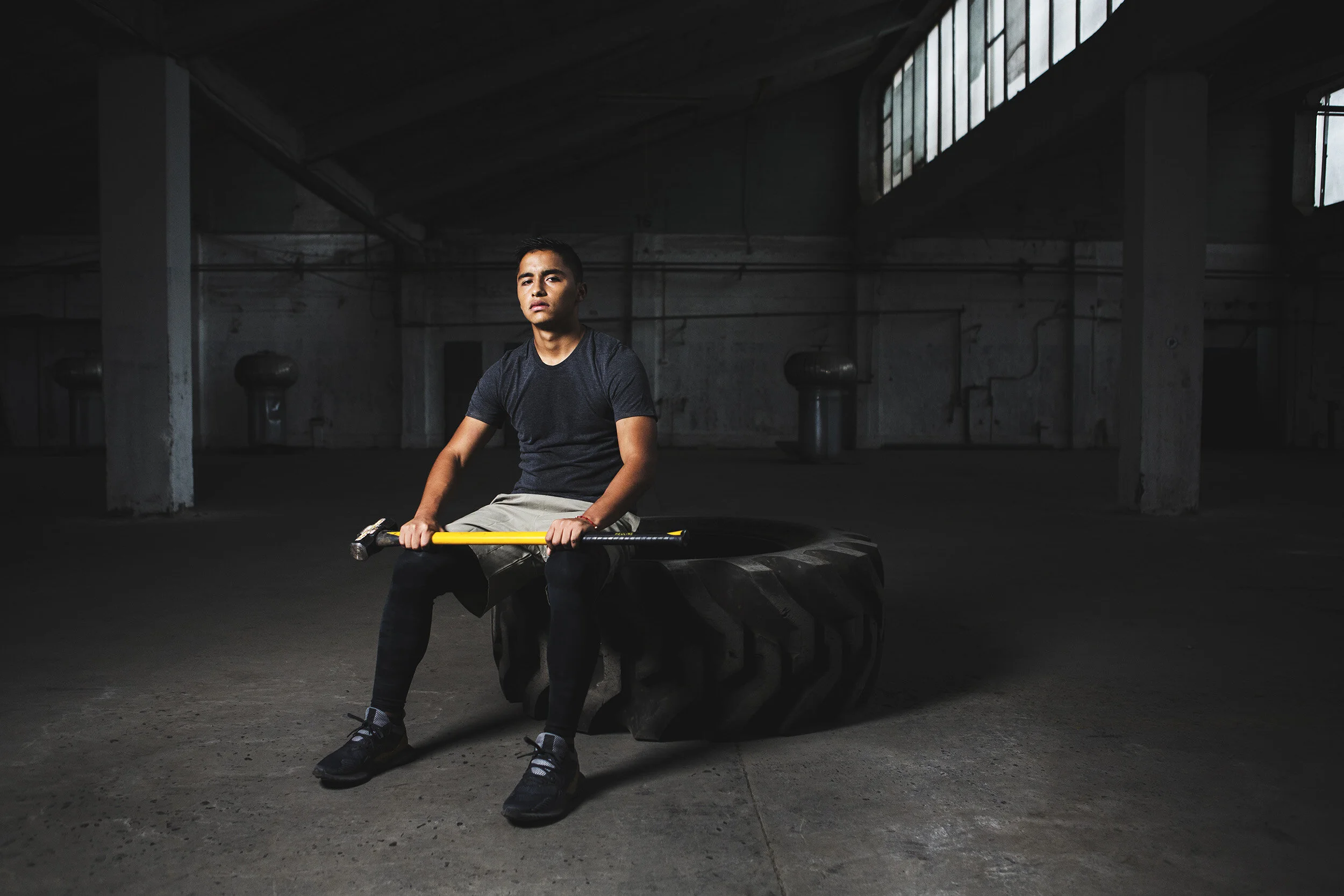 A man sitting on an industrial tire in a dimly lit, empty warehouse holding a sledgehammer with a yellow handle.