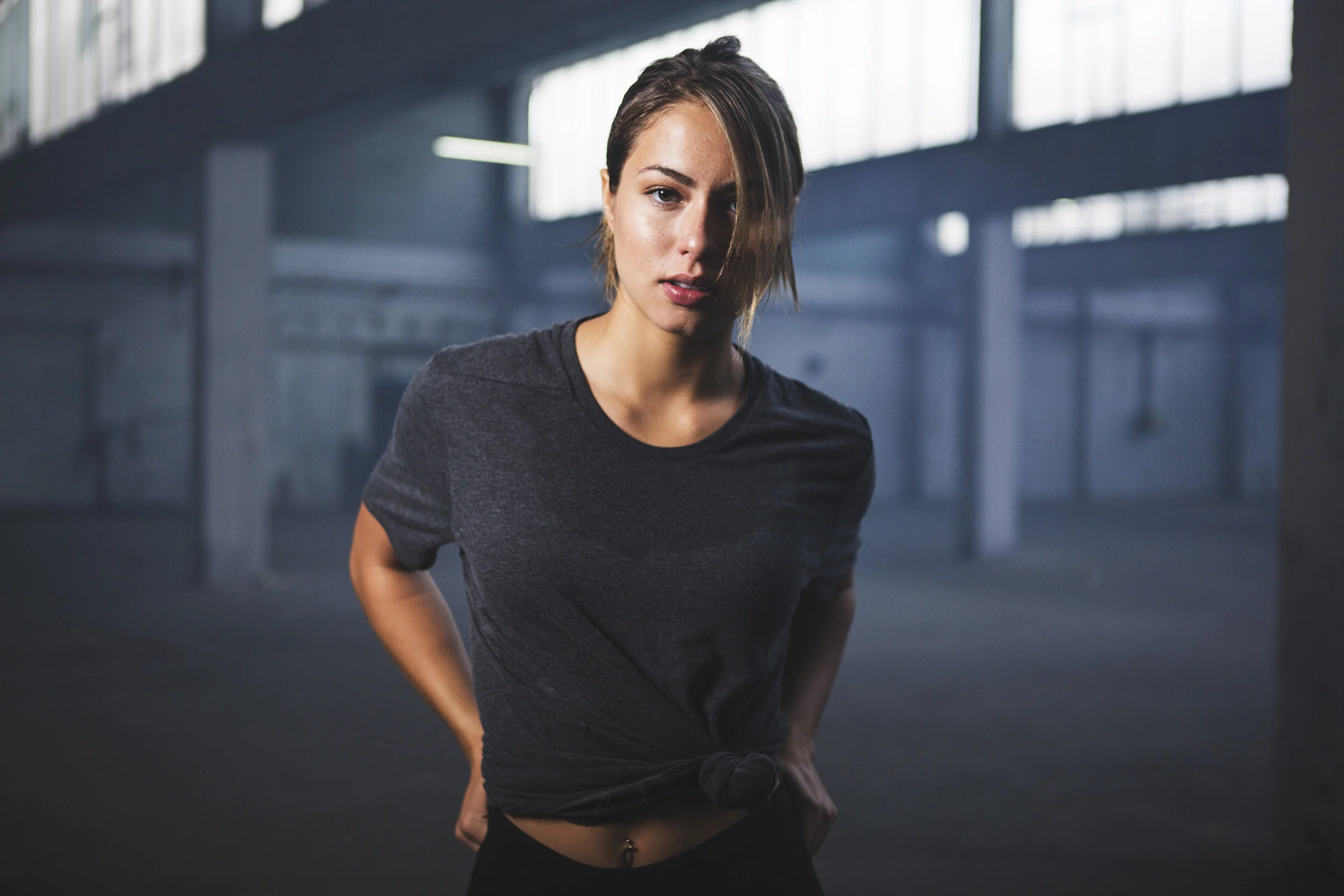 A woman with short hair in a dark t-shirt and black pants, standing in an industrial indoor space with a serious expression.
