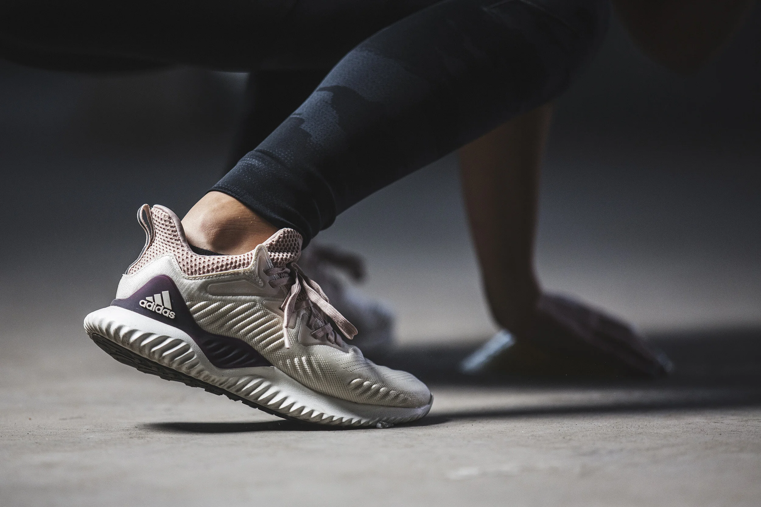 A person in black leggings and pink Adidas running shoes is in a starting position on a concrete surface, preparing to push off in a sprint or workout.