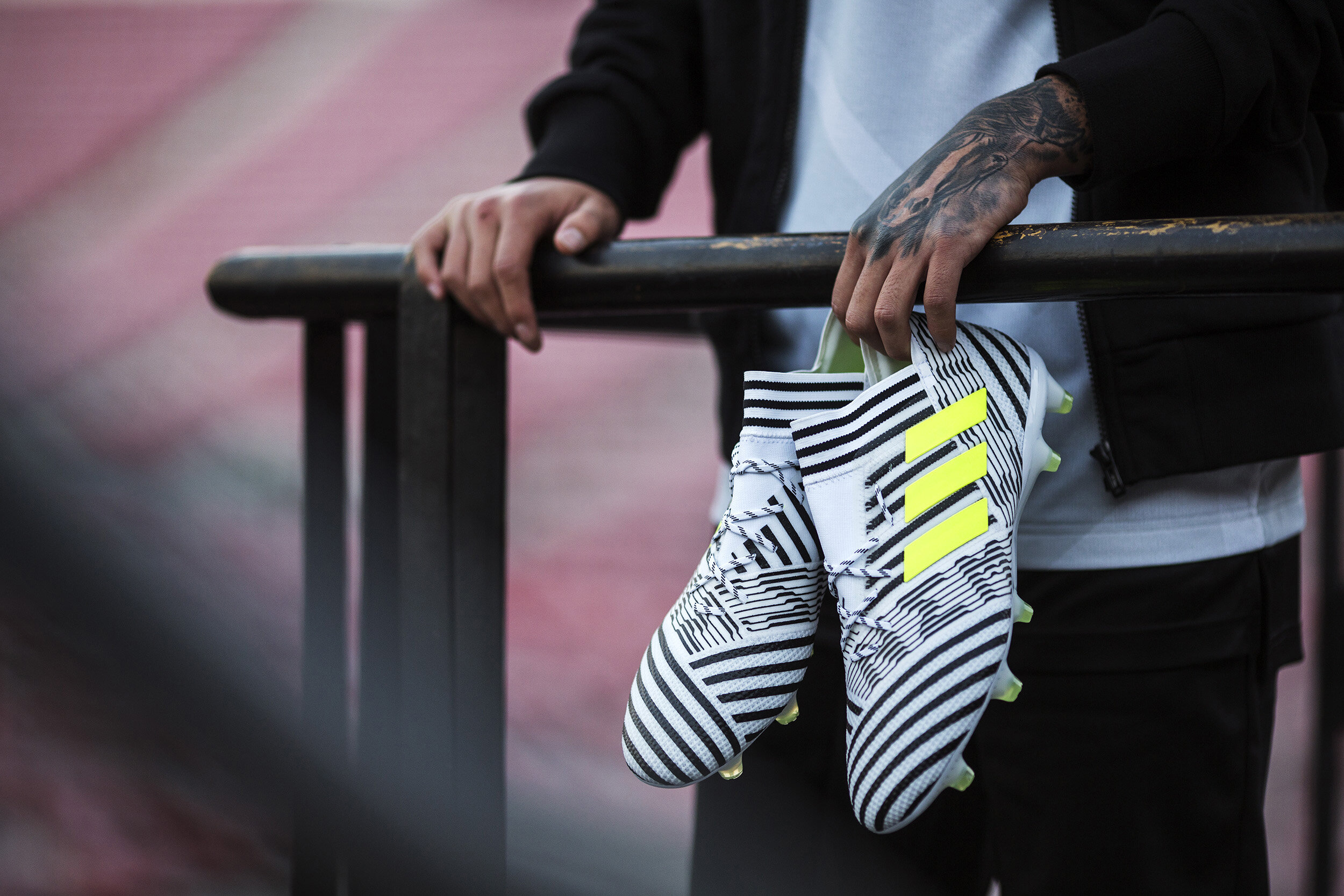 Person holding a pair of white Adidas soccer cleats with black stripes and yellow accents, leaning on a black railing.