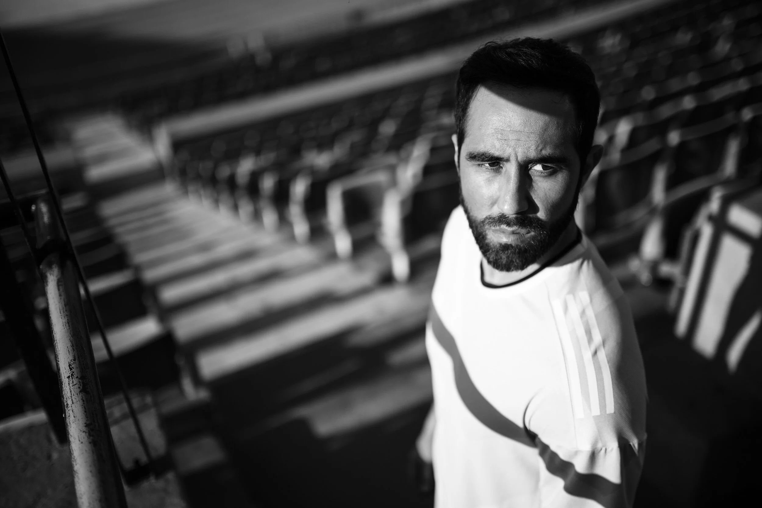A man with a beard and short hair looking to the side, wearing a white shirt with stripes on the sleeves, in an outdoor stadium with empty seats and casting shadows.