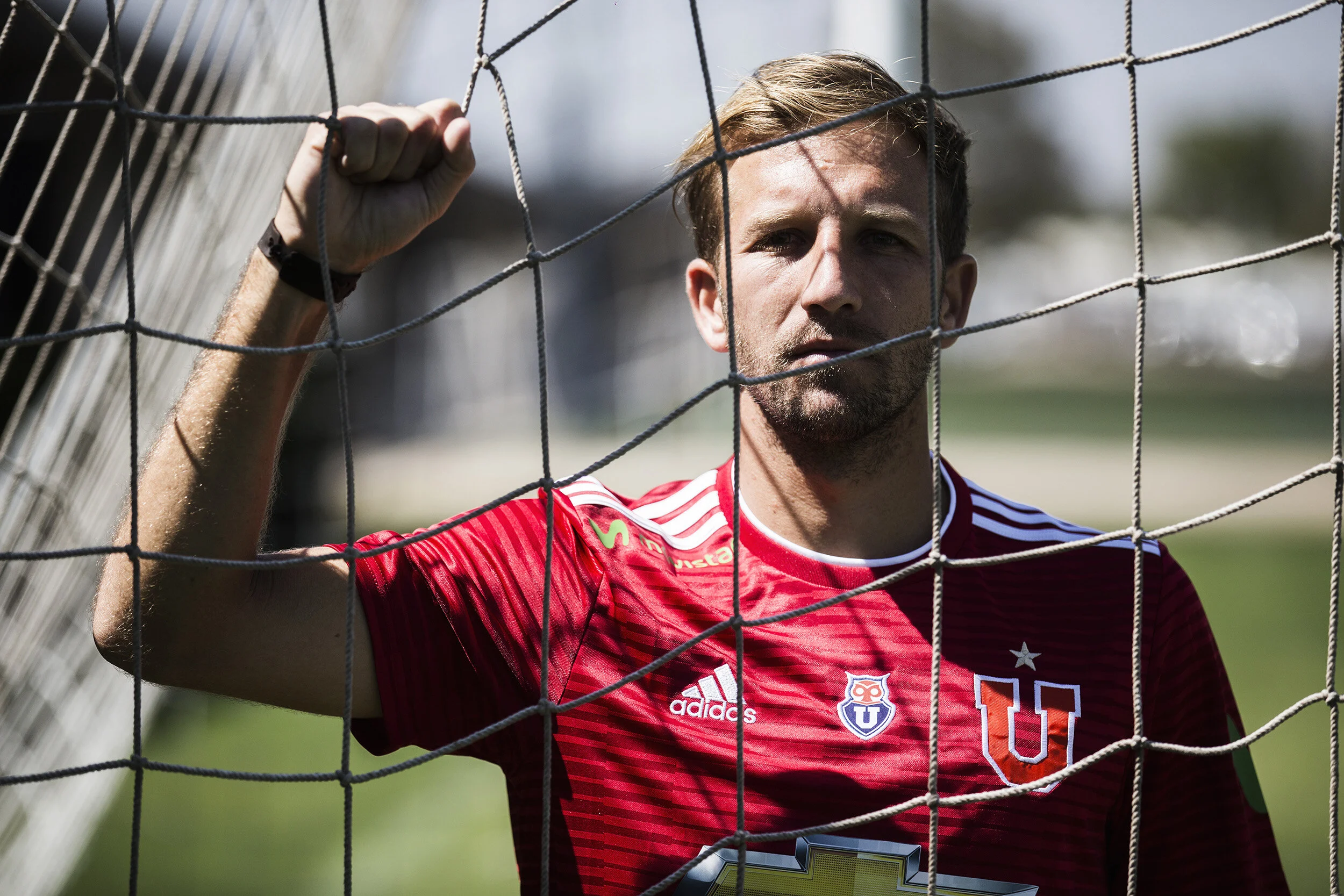 A male soccer player wearing a red jersey with U and Adidas logos, standing behind a soccer goal net.