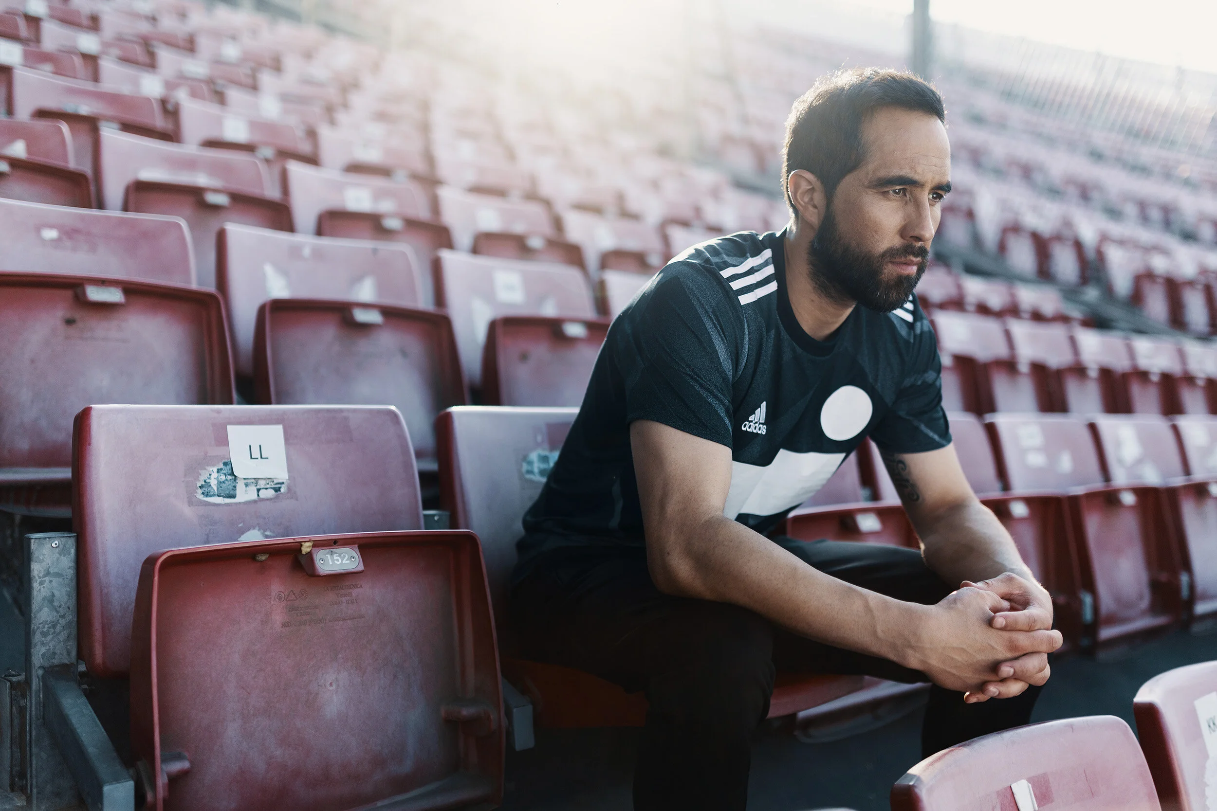 A man with a beard and dark hair sitting alone on empty stadium seats with a focused expression, wearing a black sports jersey with white accents.
