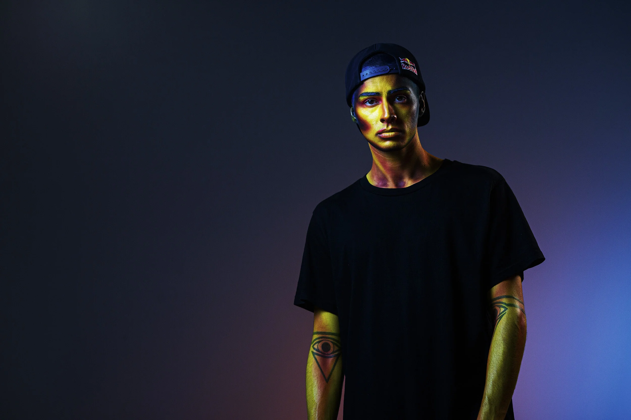 A young person with a cap backwards and a black T-shirt, with colorful face paint and tattoos, standing against a gradient dark background.