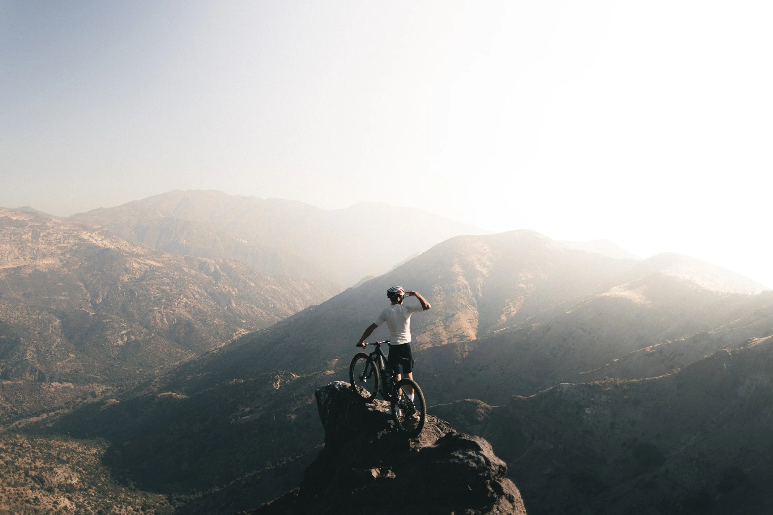A person with a helmet standing on a mountain rock with a mountain landscape in the background, holding a bicycle and looking into the distance.