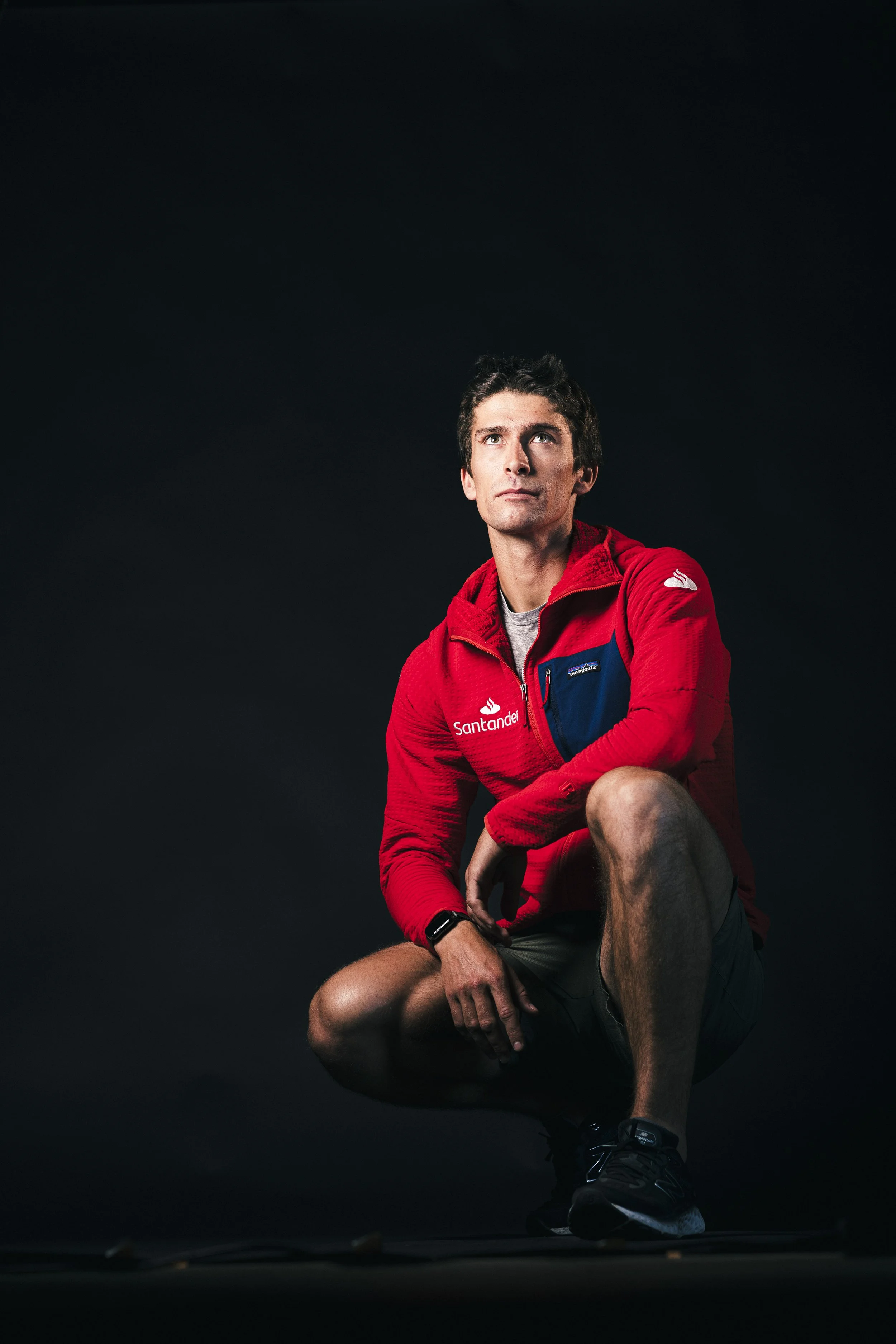 A young man crouching, looking up, wearing a red jacket with multiple sponsor logos, shorts, and running shoes, against a dark background.