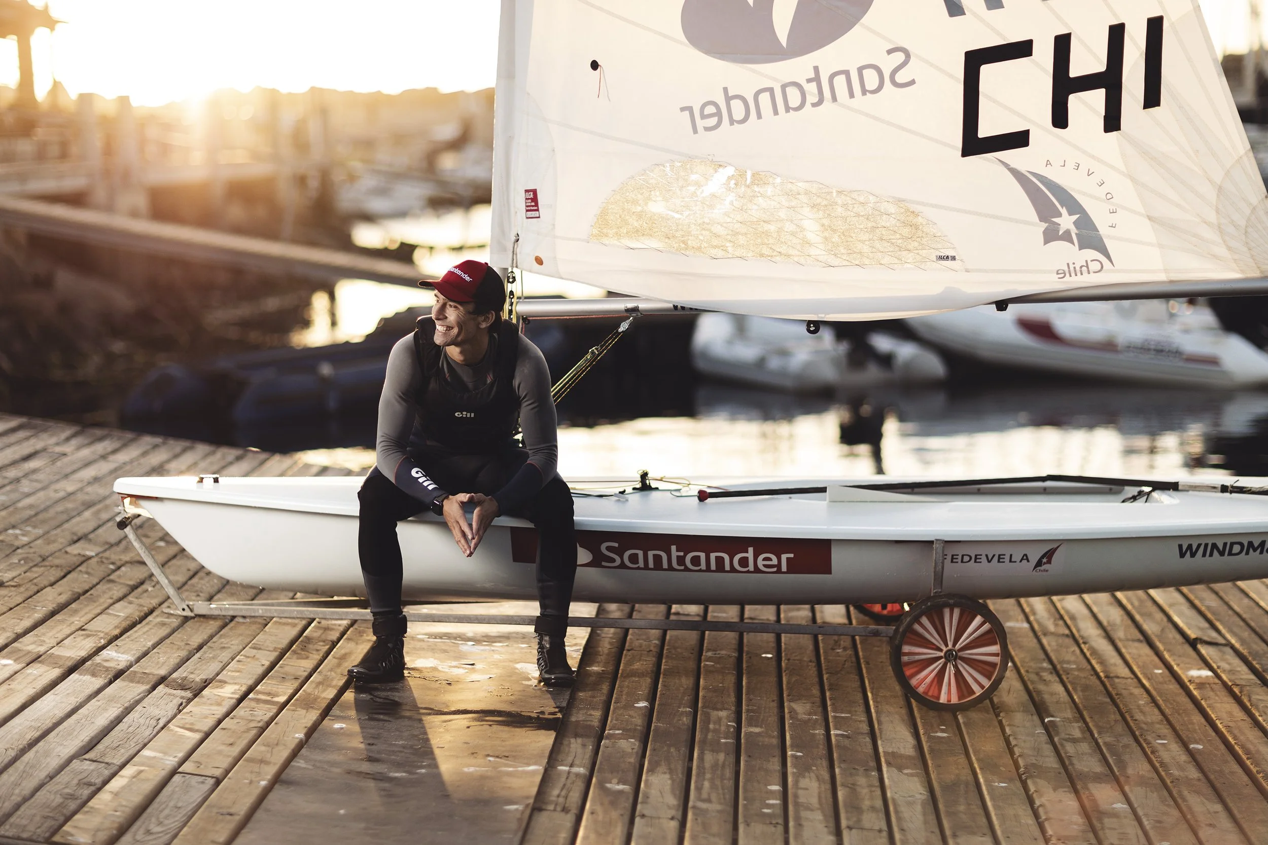 A smiling woman in sailing gear sitting on the edge of a small racing sailboat, docked on a wooden pier during sunset.