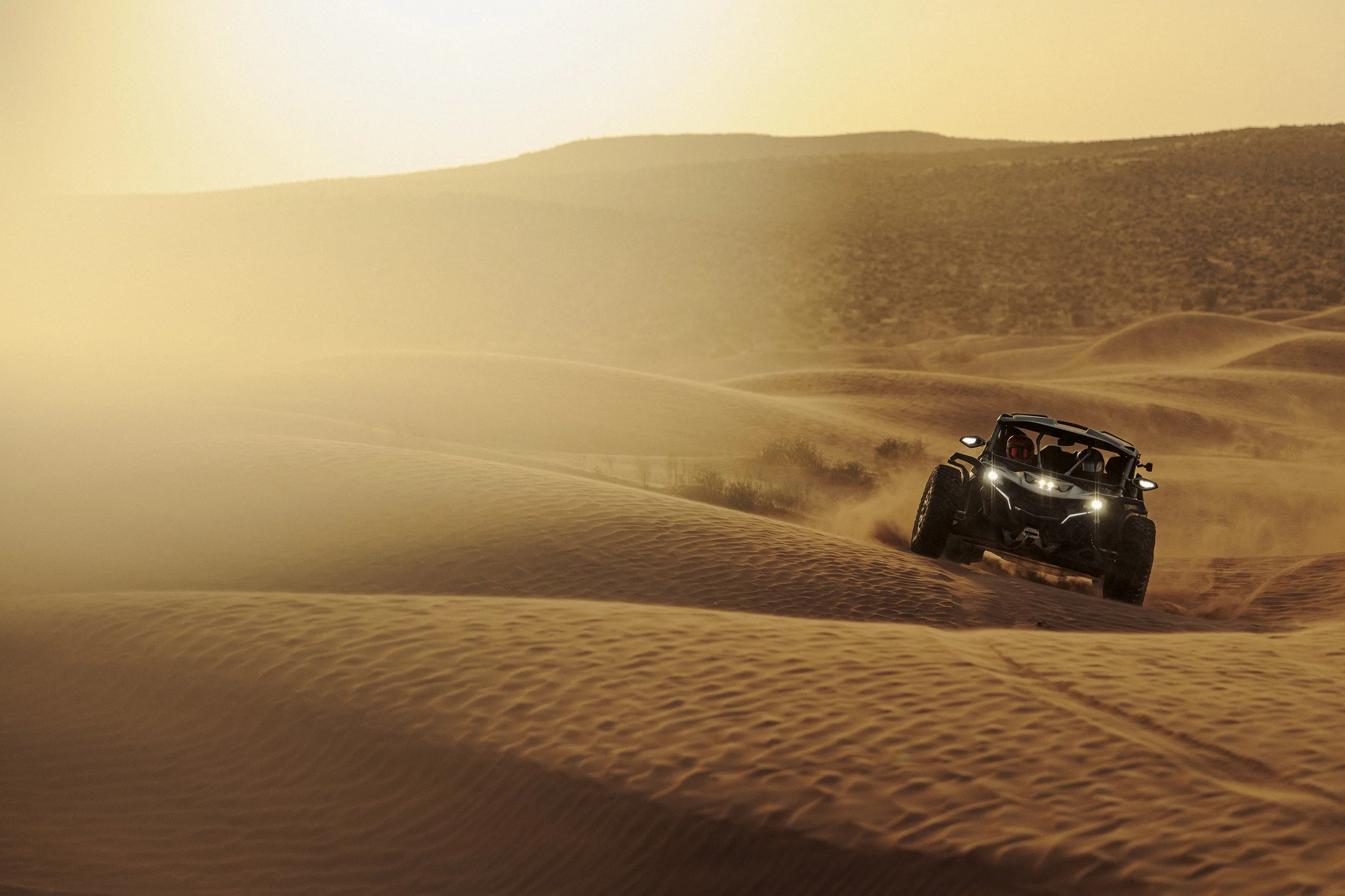 An all-terrain vehicle driving on sand dunes in a desert landscape at sunset or sunrise.