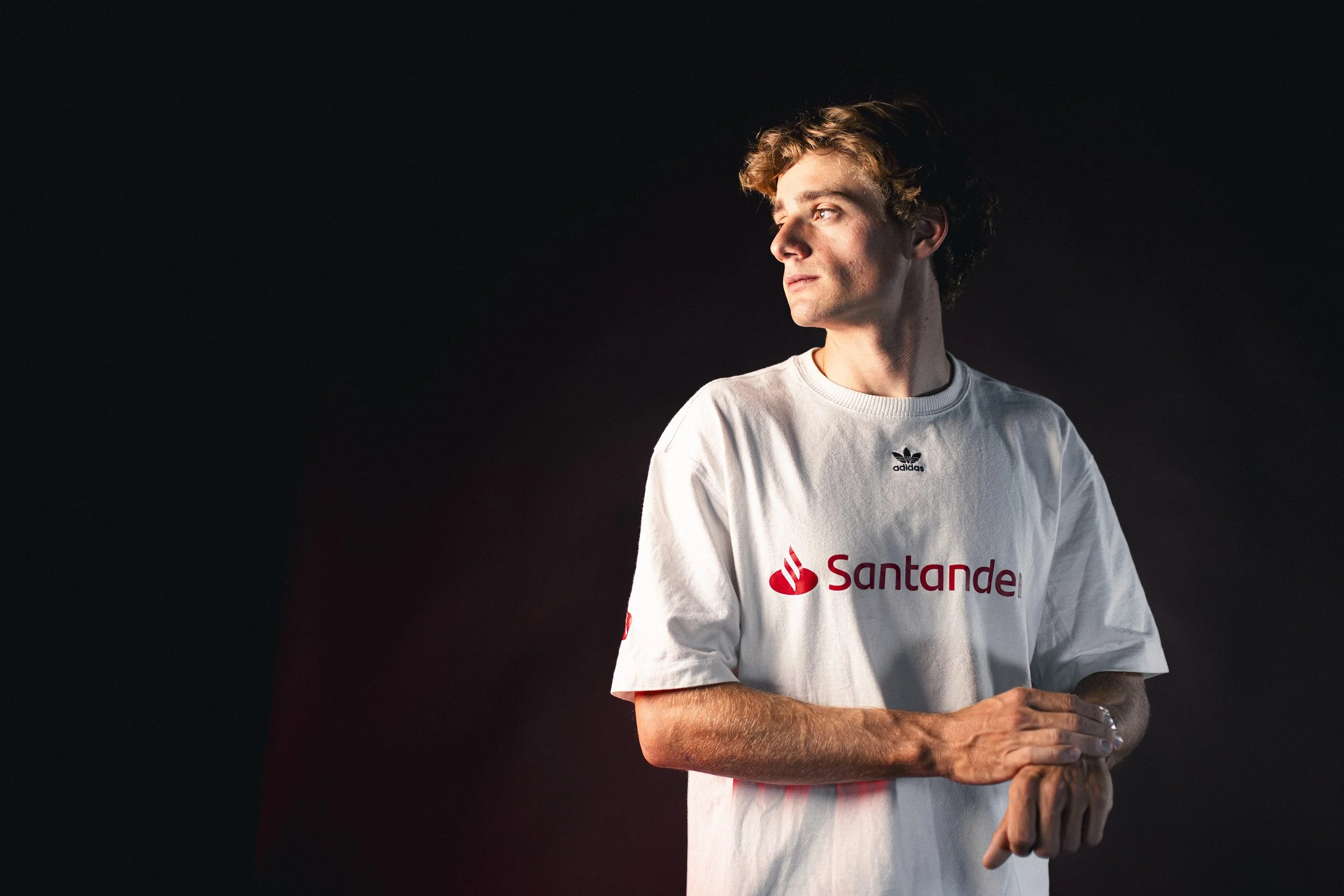 A young man with curly hair wearing a white sports jersey with 'Santander' logo and the Adidas logo, standing against a dark background, looking to his left with a thoughtful expression.