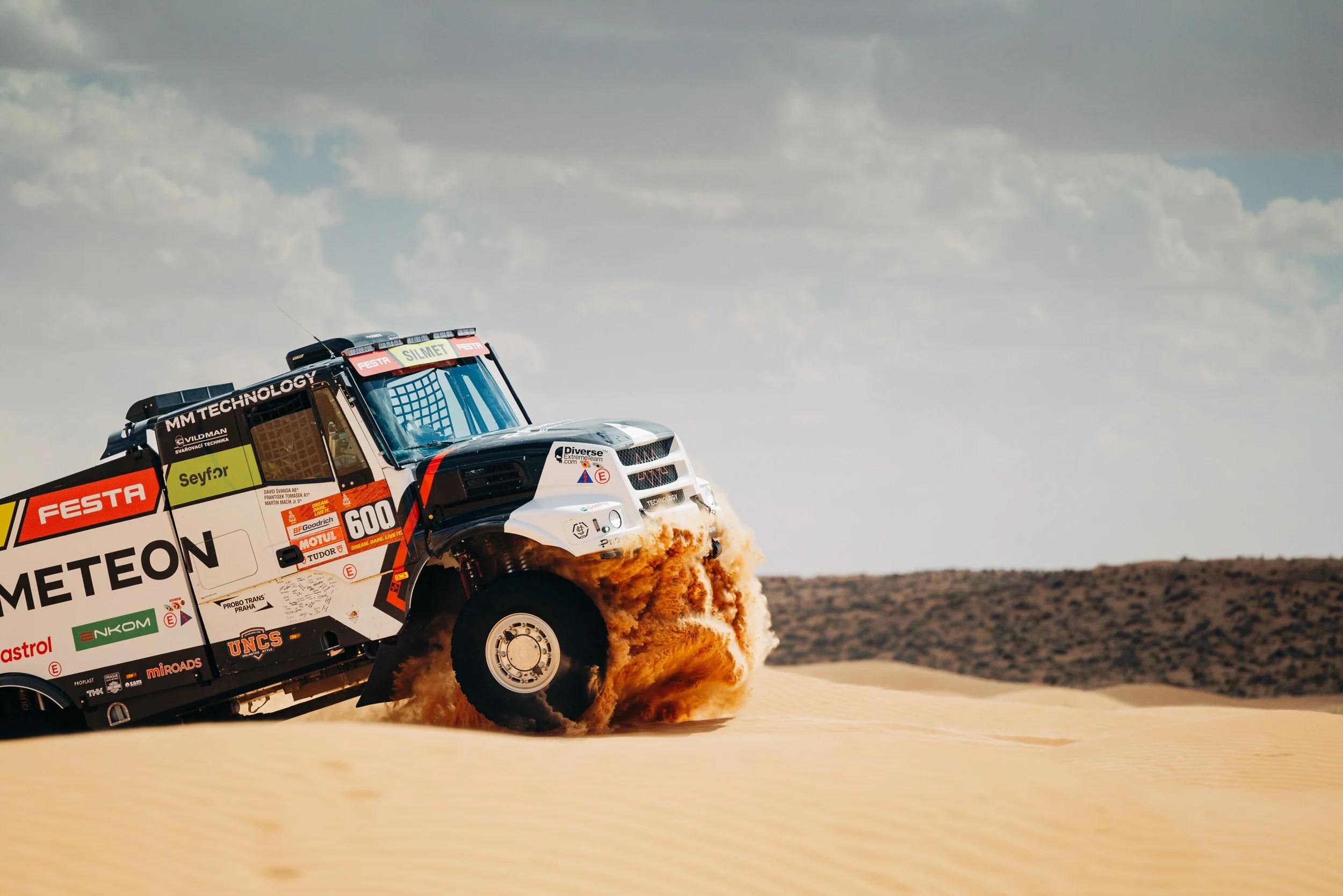 A rally truck kicks up dust in a desert landscape, with a cloudy sky overhead.