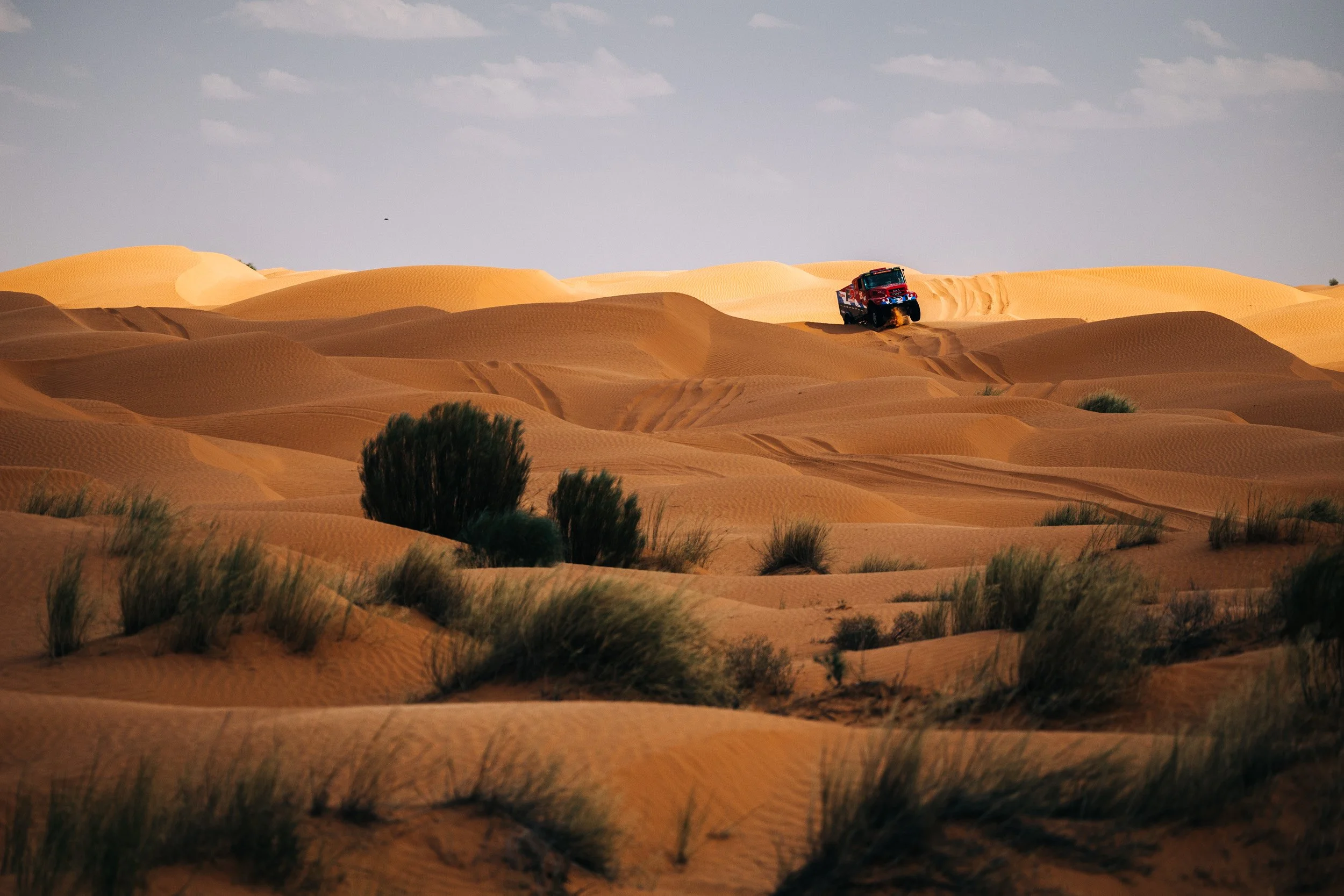 A red off-road truck driving through sand dunes in a desert landscape under a partly cloudy sky.