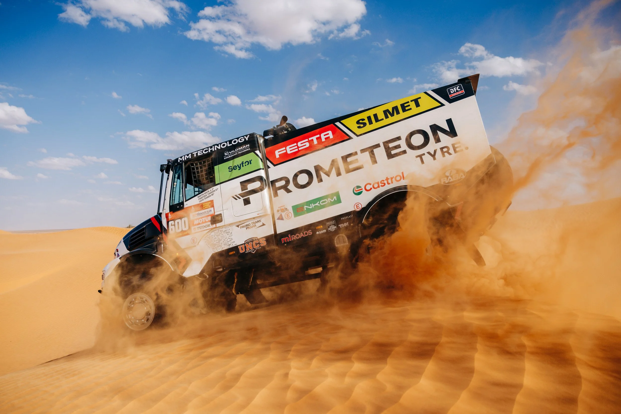 A rally truck kicking up dust as it drives across a sandy desert landscape under a blue sky with scattered clouds.
