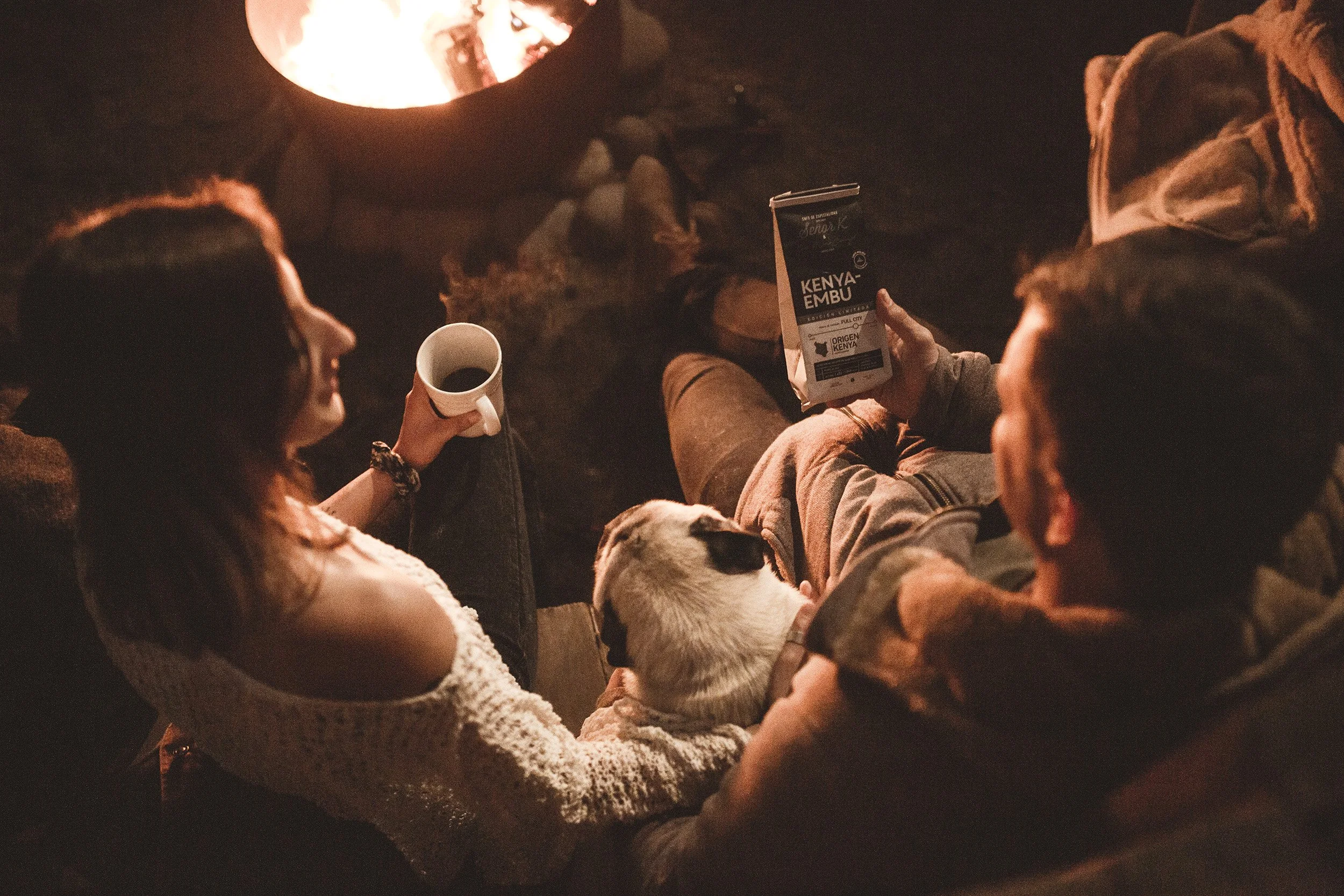 Two people, a woman and a man, sitting by a campfire at night, with a dog resting on the woman's lap. The woman holds a mug, and the man holds a package of Kenyan coffee.