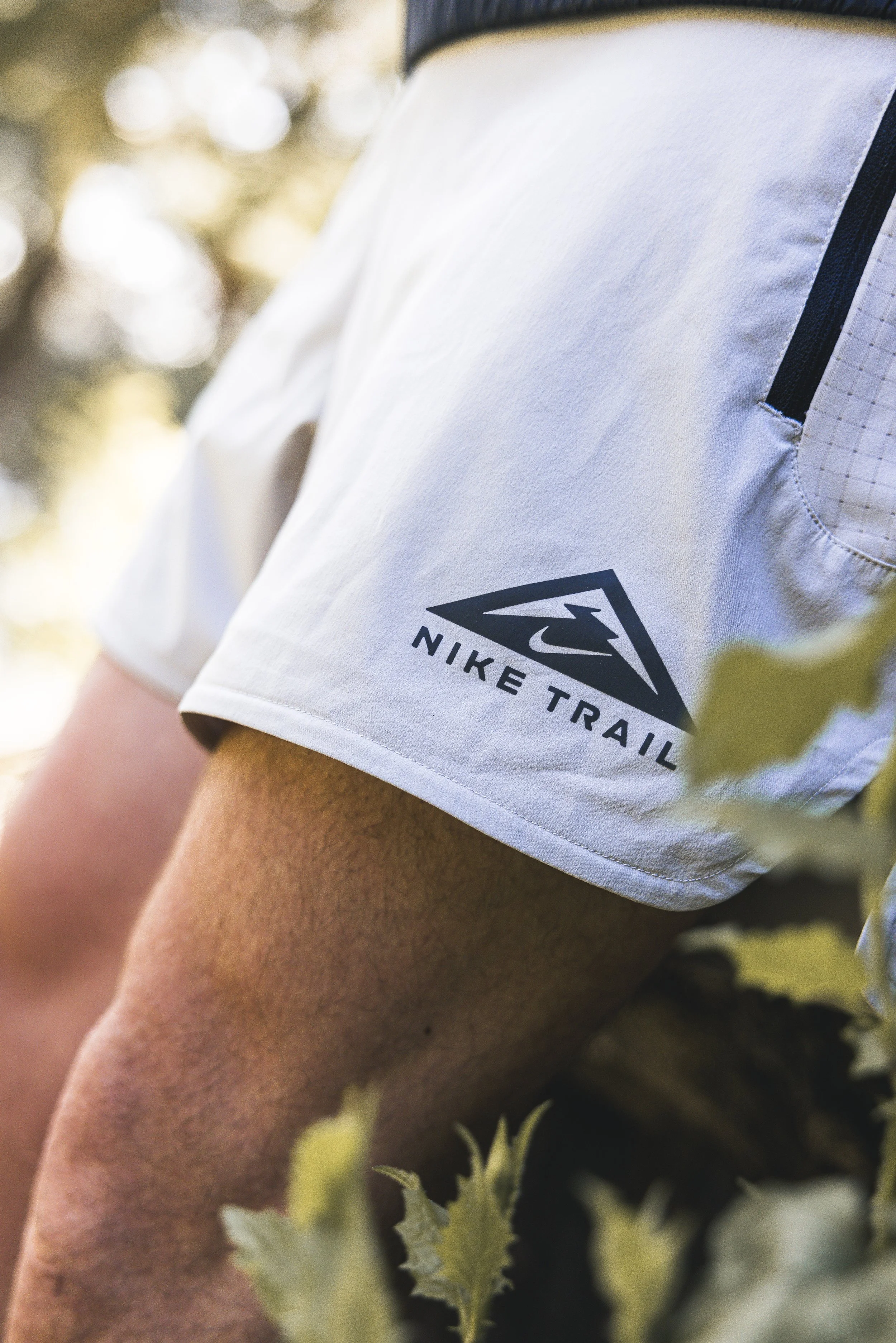 Close-up of a person wearing a white Nike Trail t-shirt, with a focus on the logo and part of the person's arm and leg, outdoors in a natural setting with blurred foliage in the background.