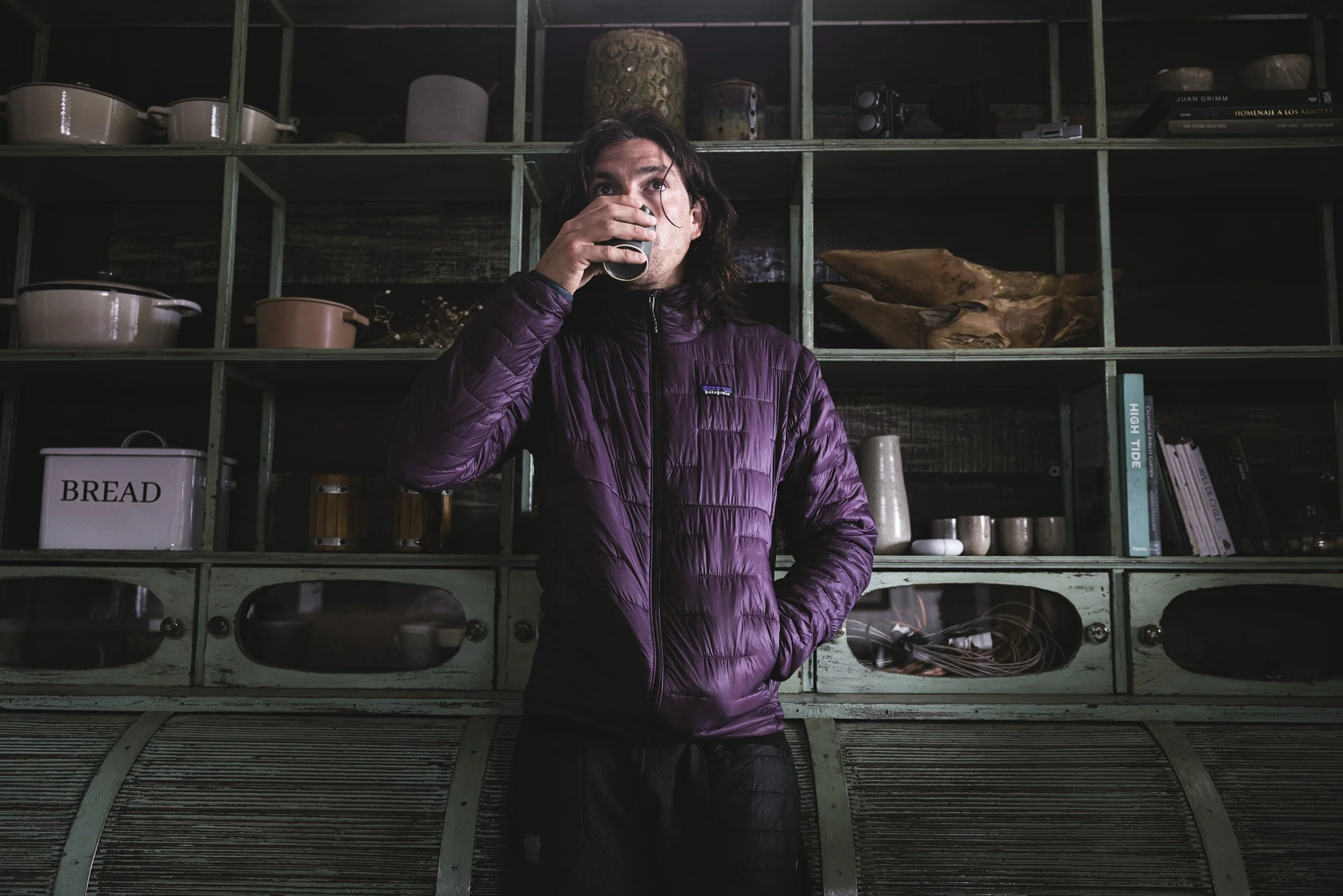 A man with long dark hair wearing a purple jacket is taking a sip from a glass of dark liquid in front of a green wooden shelf filled with various kitchen items and books.