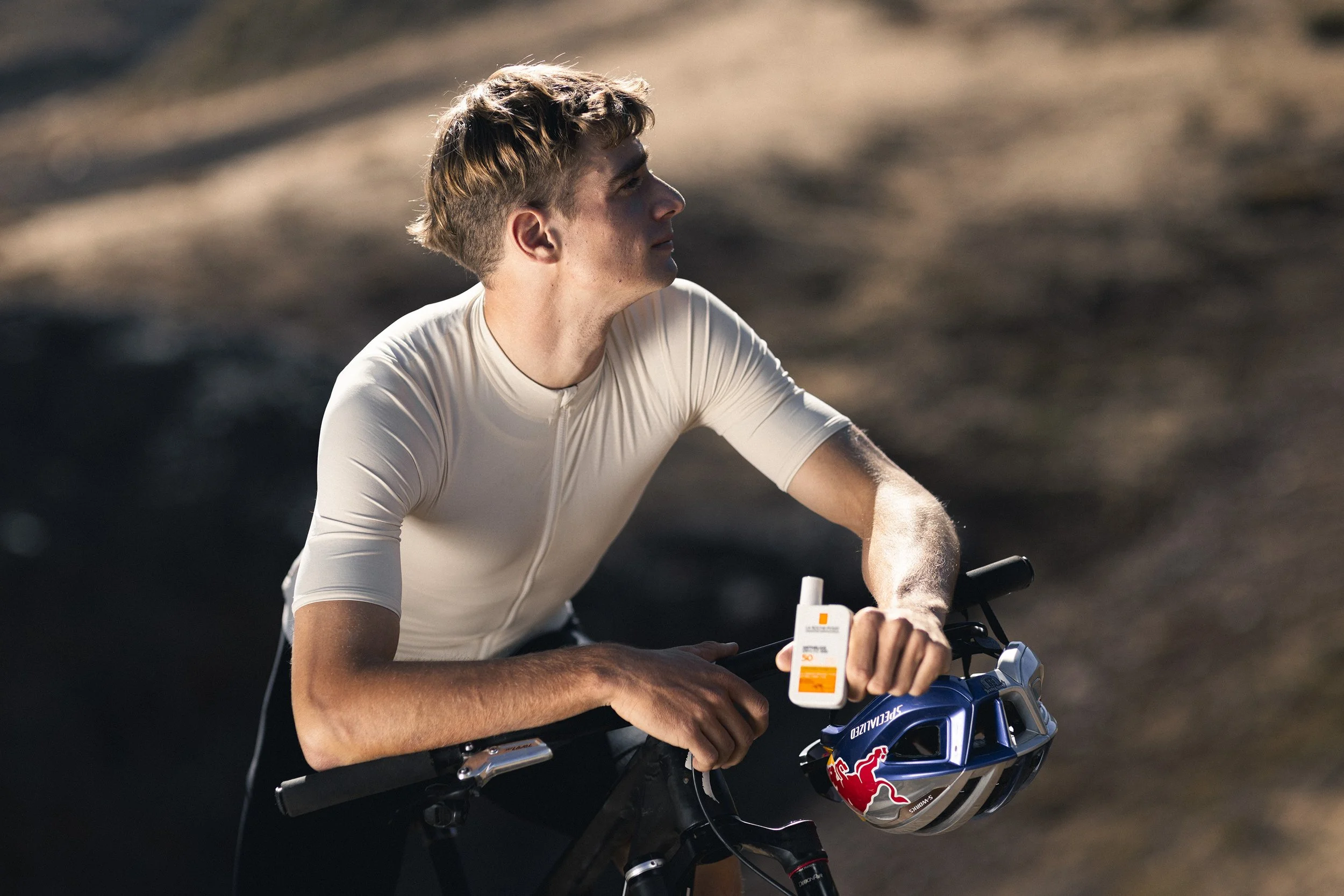 A young man in a white athletic shirt resting on a bicycle handlebar, holding a tube of sunscreen, with a red and blue helmet hanging on the bike's handlebar, outdoors with a blurry background.