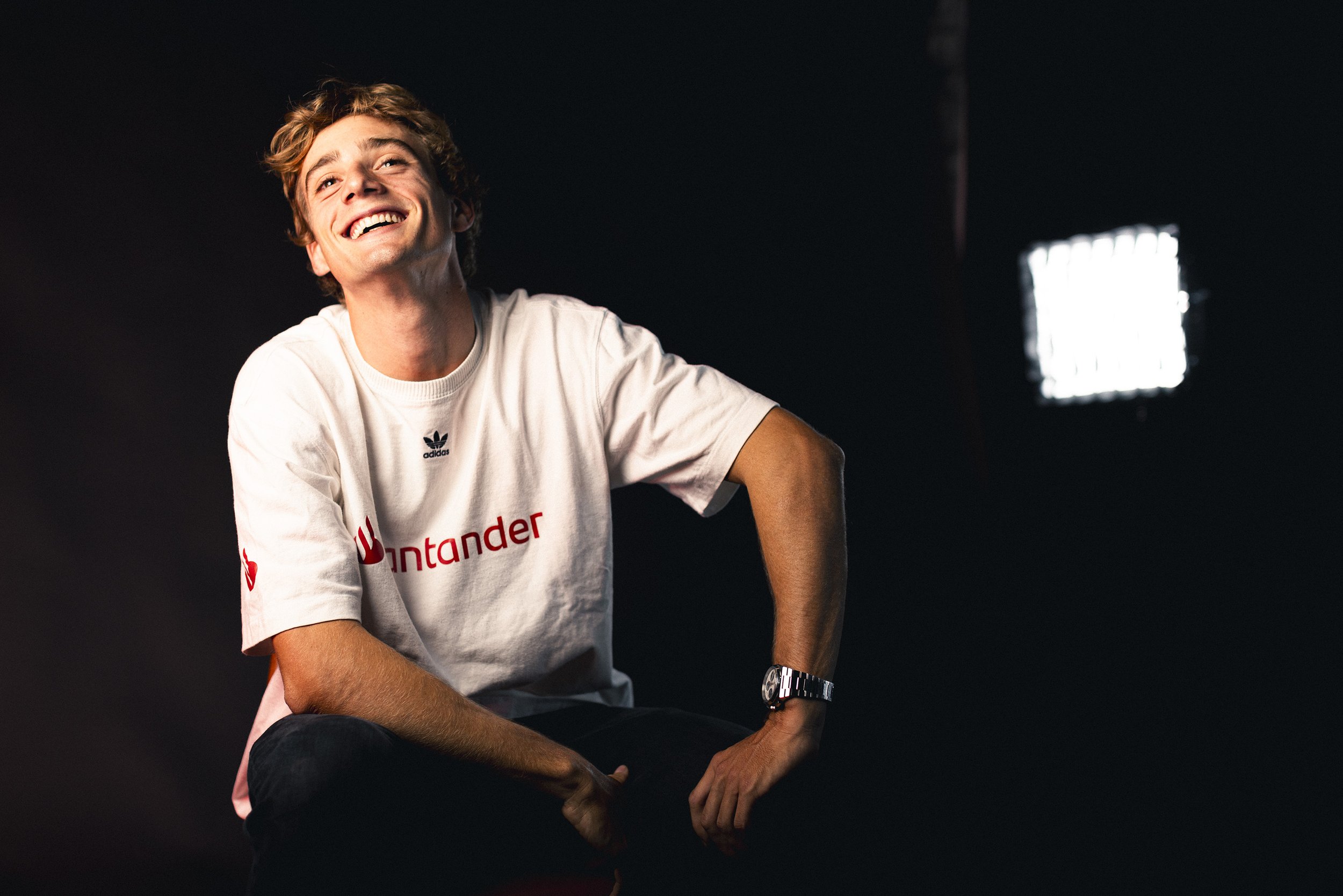 A young man with brown curly hair is smiling and appears happy. He is wearing a white t-shirt with red and black logos, including 'Santander' and Adidas, a silver wristwatch, and dark pants. The background is dark with a bright light source in the di