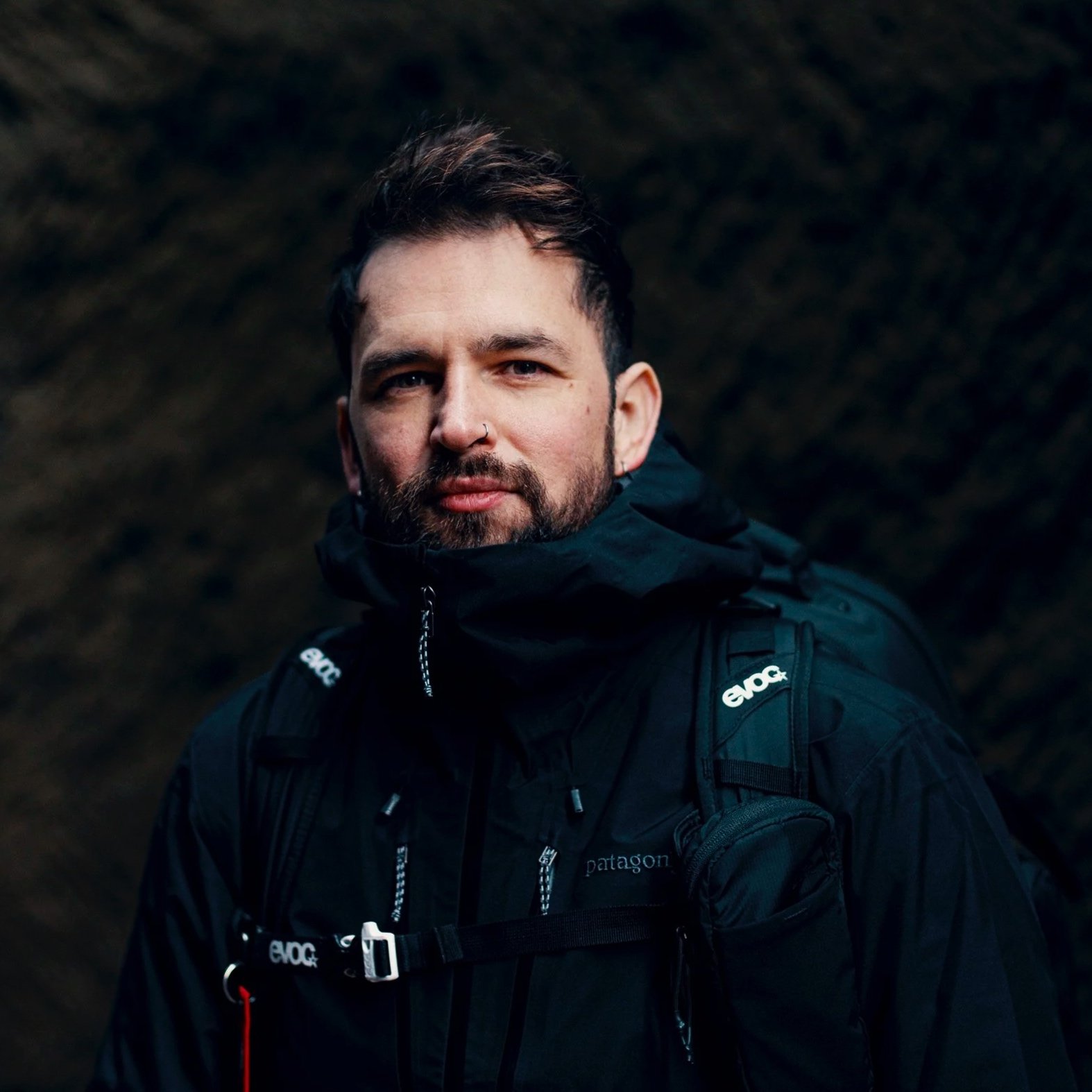 A man with short brown hair and a beard, wearing a black outdoor jacket and backpack, standing in a dimly lit cave or dark environment.