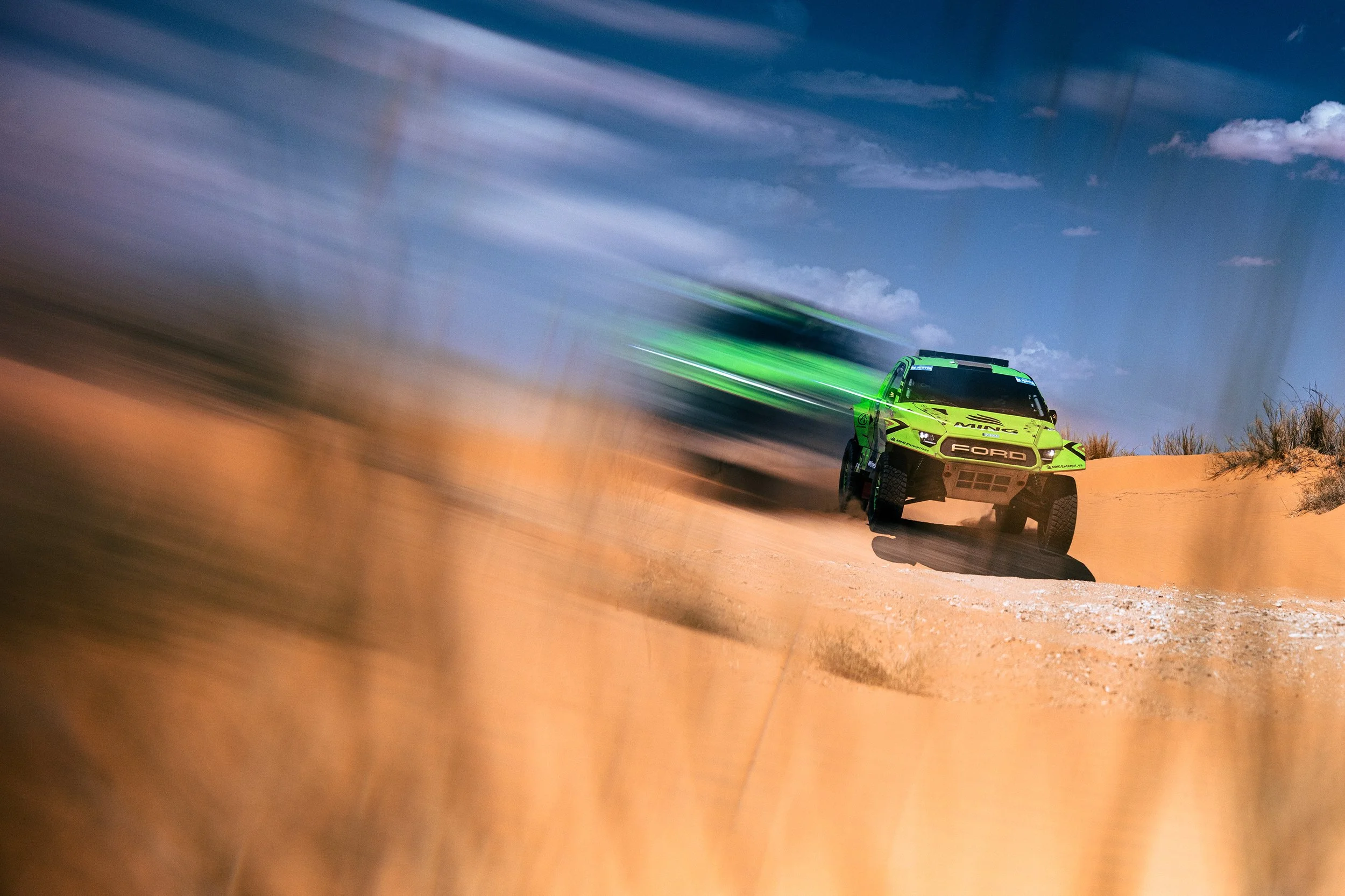 A green and black Ford rally car speeding through a sandy desert landscape with a blue sky and scattered clouds, creating a motion blur effect.