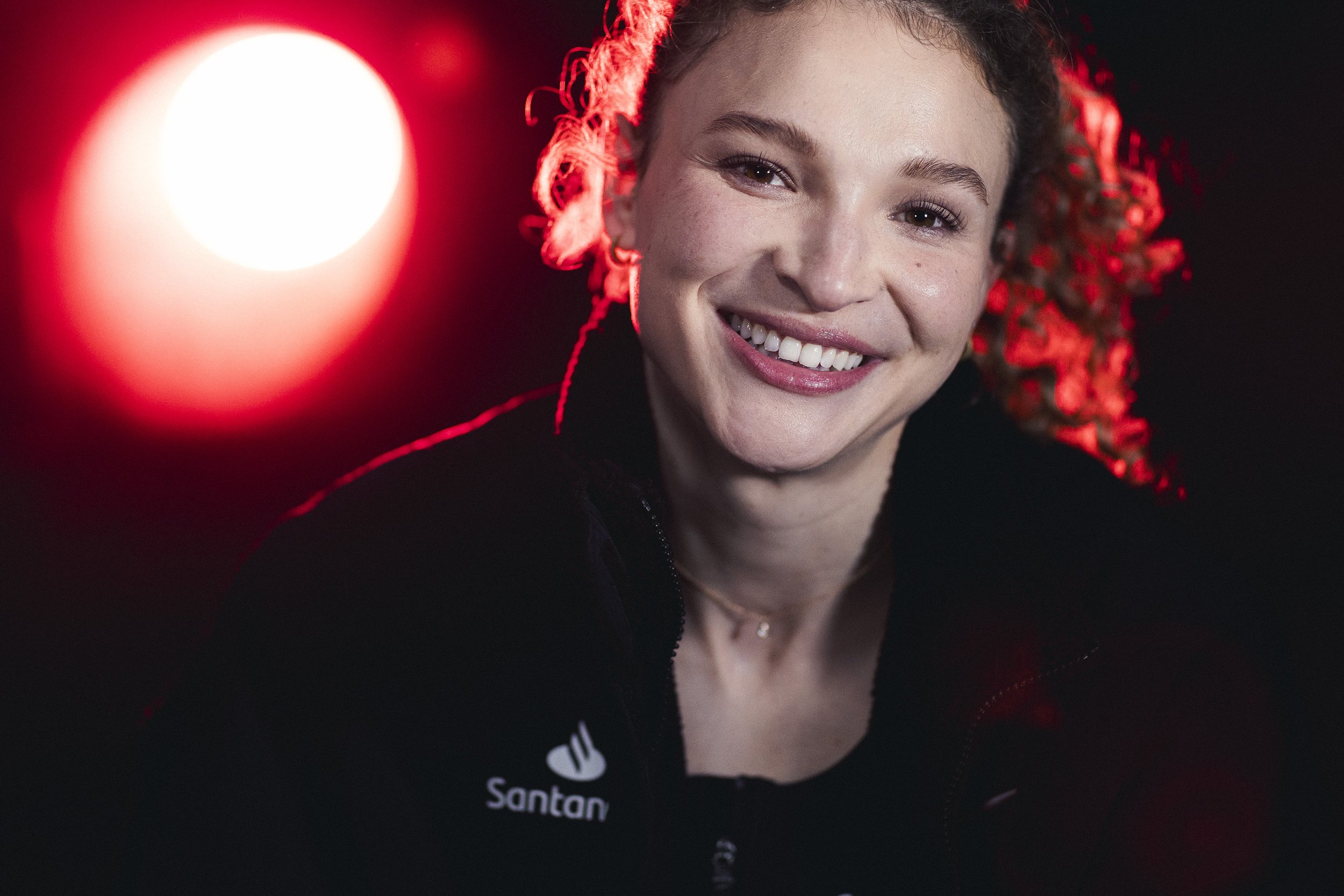 Smiling woman with curly hair, wearing a black jacket with a 'Santan' logo, illuminated by red and white lights in the background.