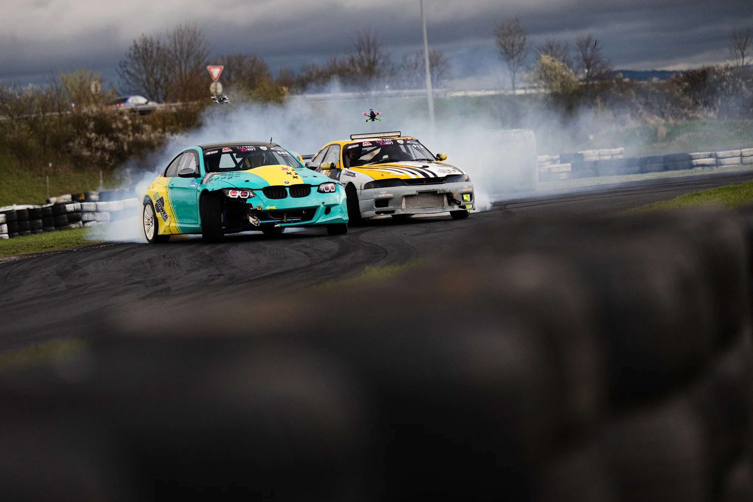 Two race cars drifting on a track, with tire smoke and remote-controlled drones flying above, under dark cloudy skies.