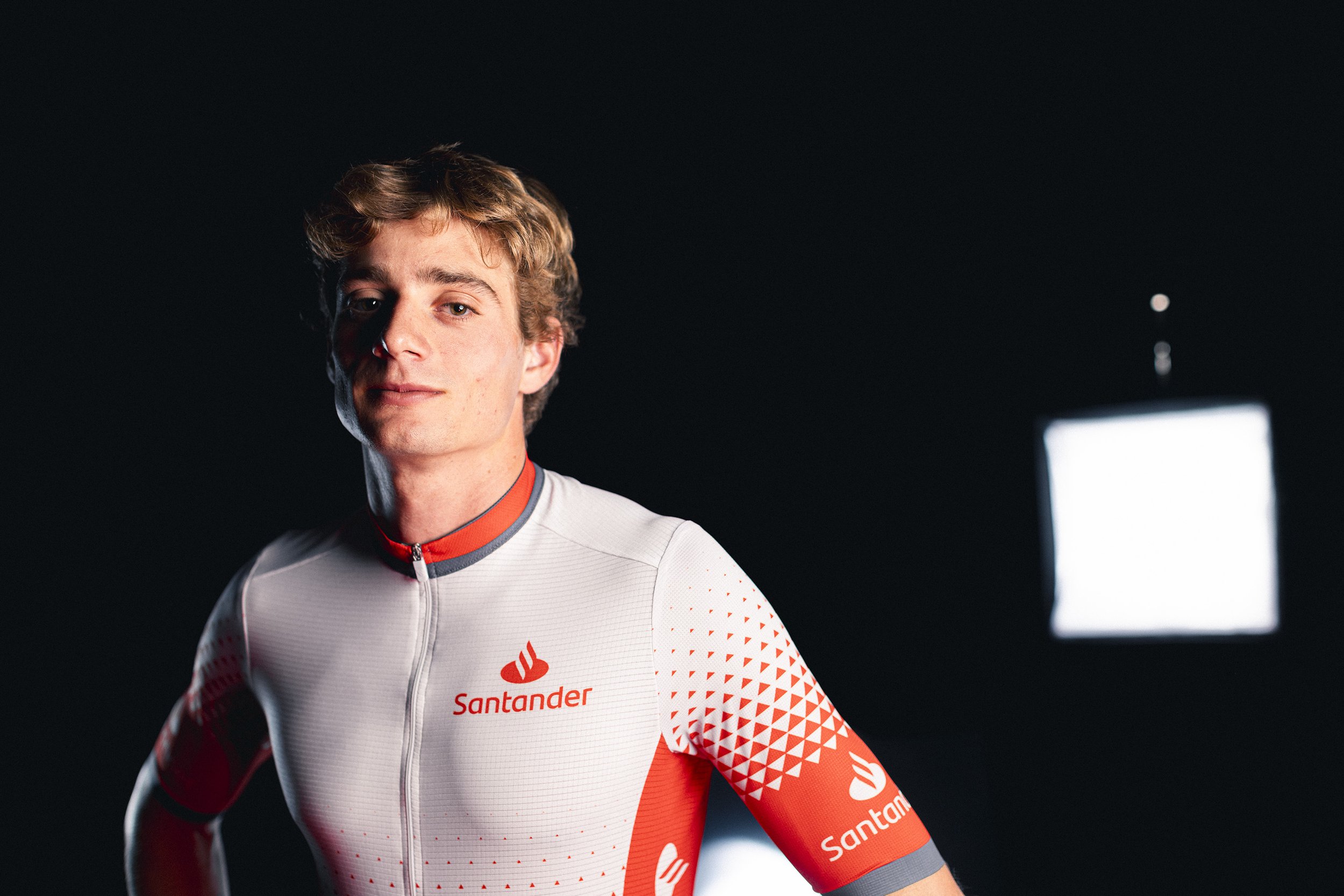 A young male cyclist wearing a white and red Santander team jersey, posing against a dark background with studio lights.