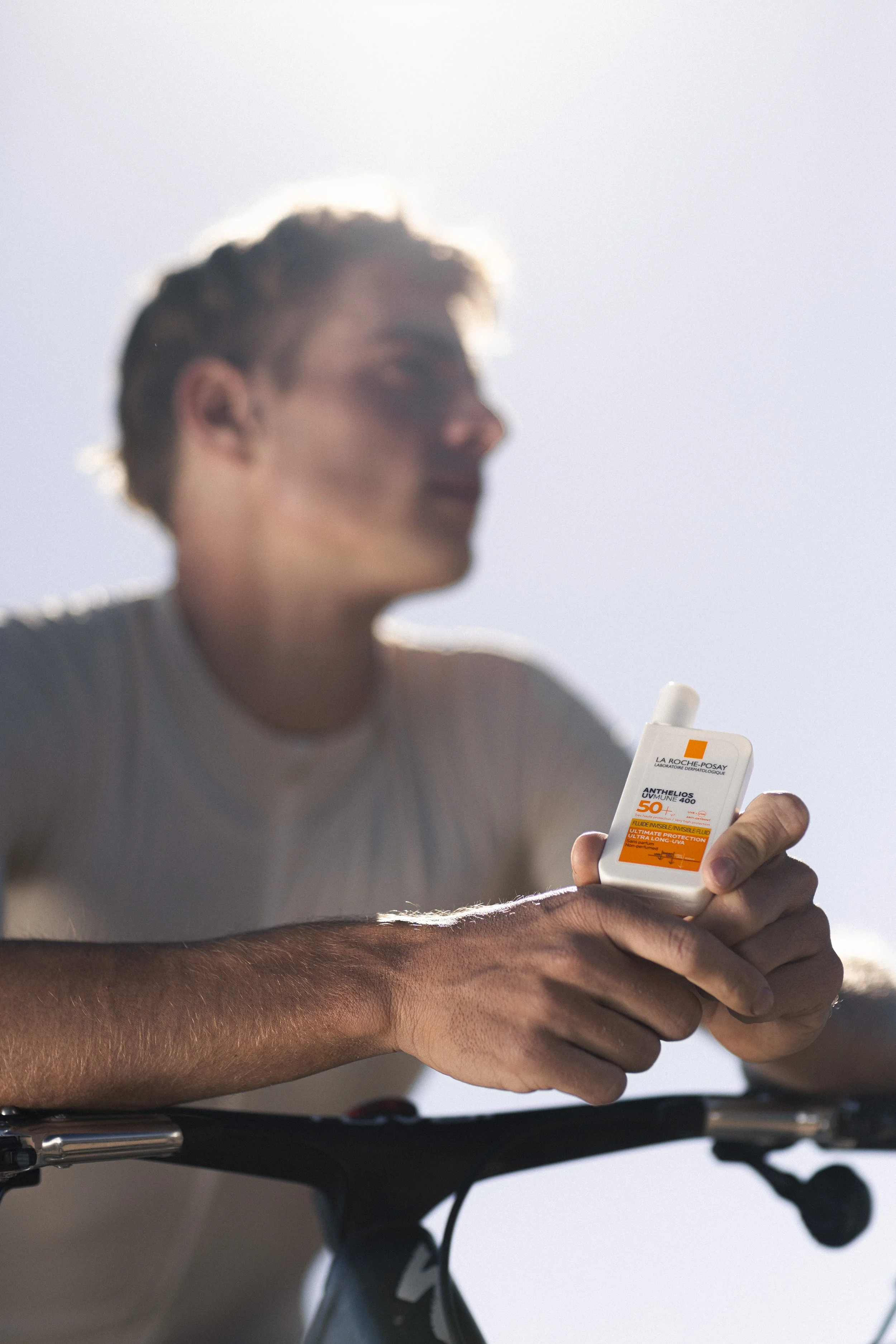 A person holding a bottle of sunscreen while sitting on a bicycle, with a bright, sunny sky in the background.