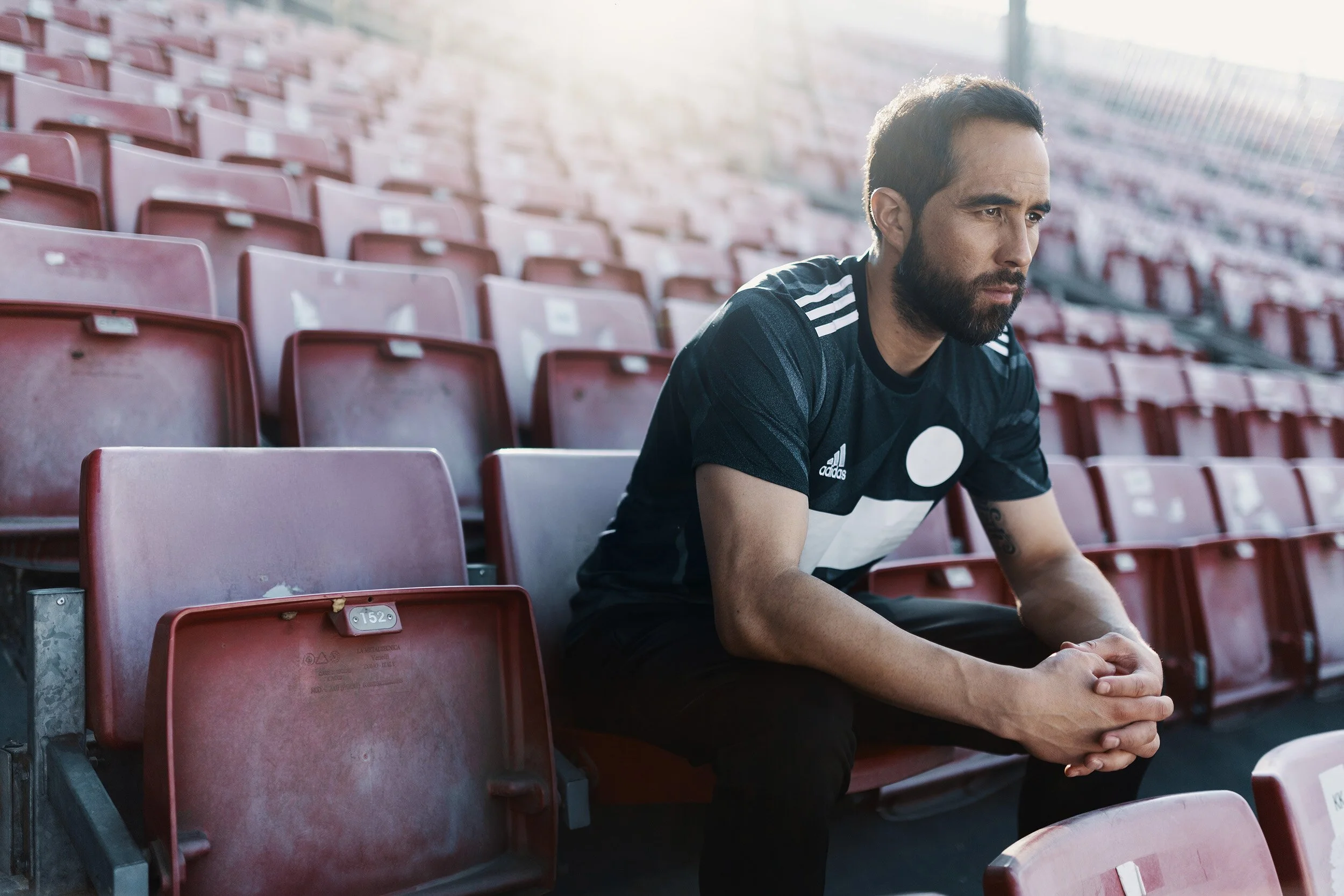 A man with a beard wearing a black Adidas T-shirt with white stripes sitting alone on red stadium seats, looking thoughtful.
