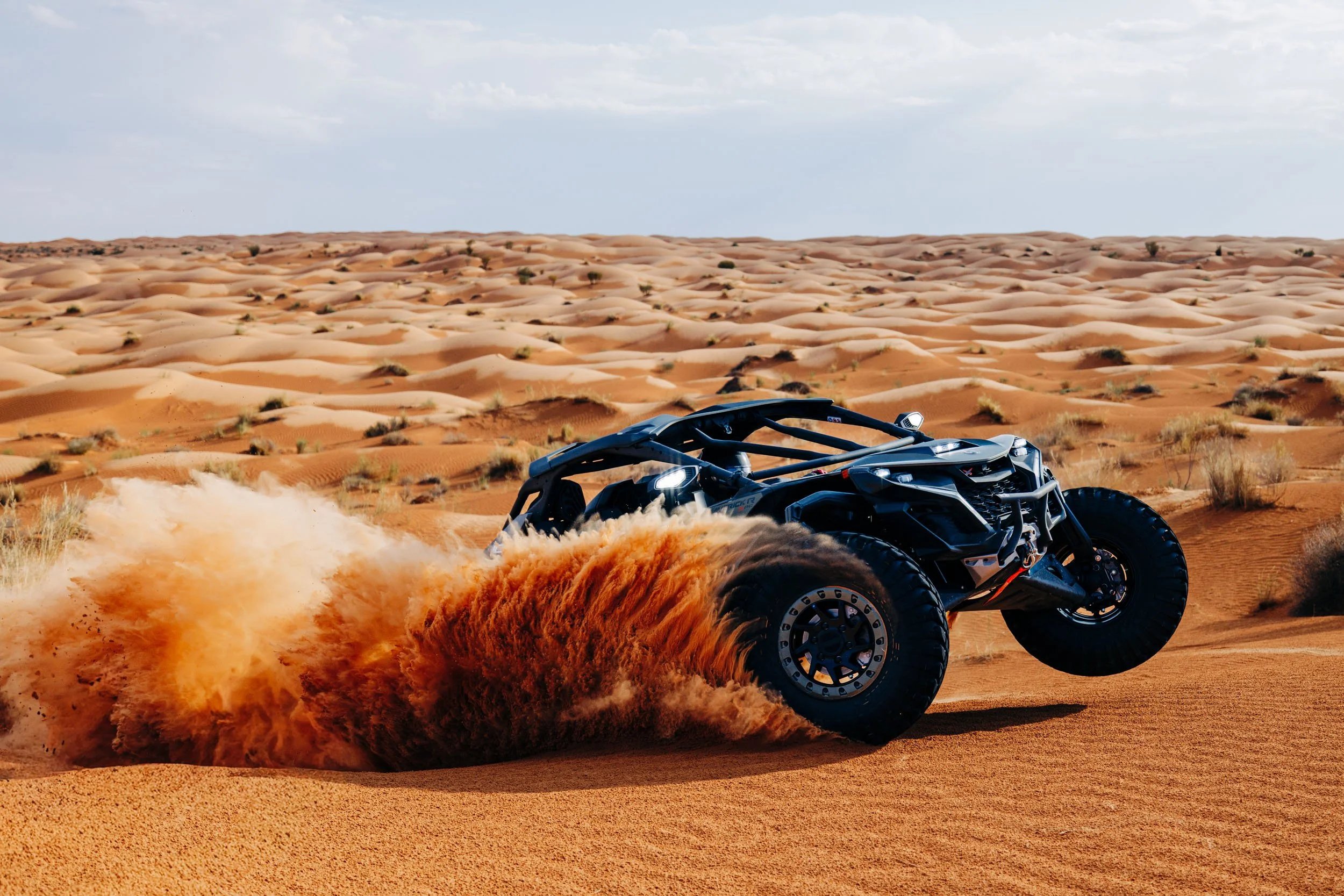 A black off-road vehicle kicking up dust on a desert landscape with sand dunes and sparse vegetation.