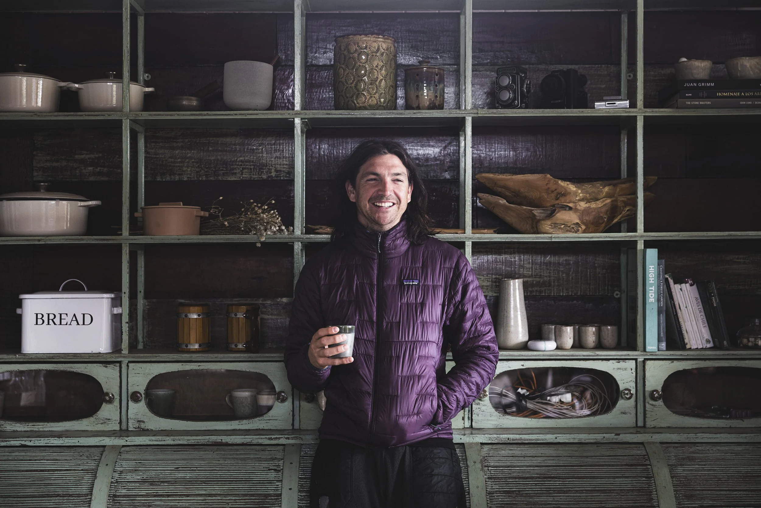 A man with long dark hair, smiling, wearing a purple puffer jacket, holding a small cup, standing in front of a green cabinet filled with kitchenware and books.