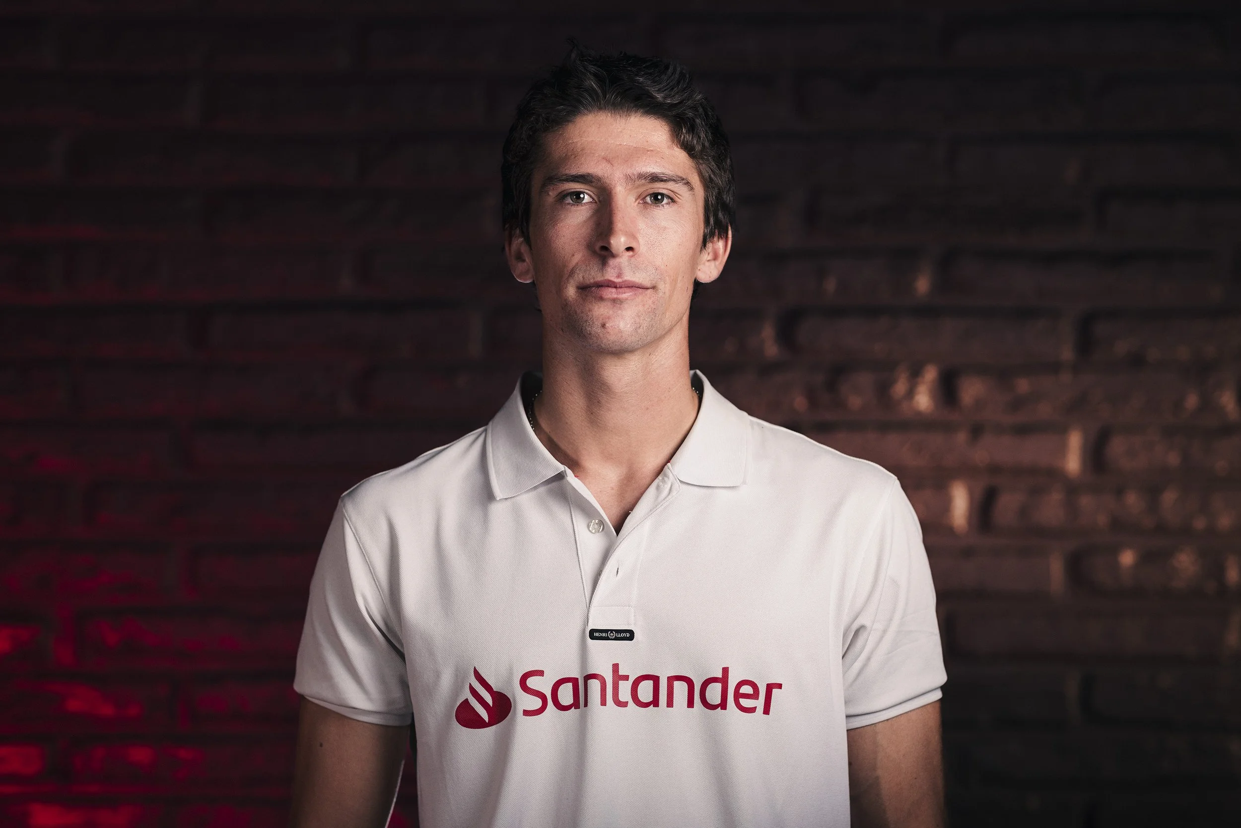 A man with dark hair wearing a white polo shirt with the Santander logo, standing in front of a dark brick wall.