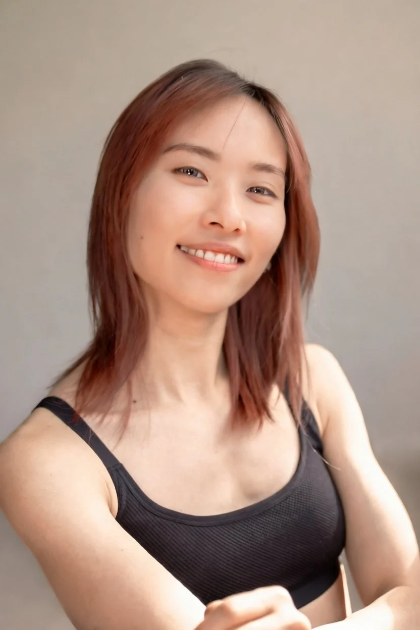 A woman who is a dance teacher at Arc Dance school in Grey Lynn, Auckland, with shoulder-length, reddish-brown hair smiling at the camera, wearing a black tank top, against a plain gray background.