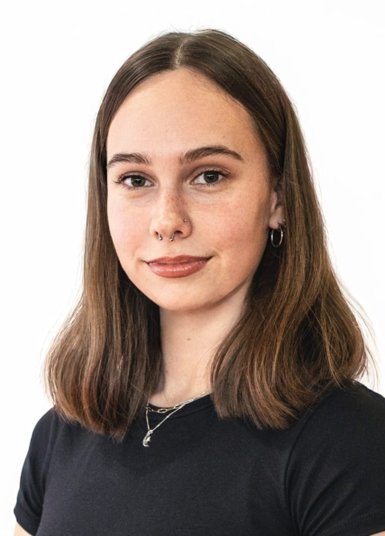 A young woman who is a dance teacher at Arc Dance school in Grey Lynn, Auckland, with brown hair, light skin, and brown eyes, wearing earrings, a nose ring, and a necklace, looking at the camera with a slight smile against a plain white background.