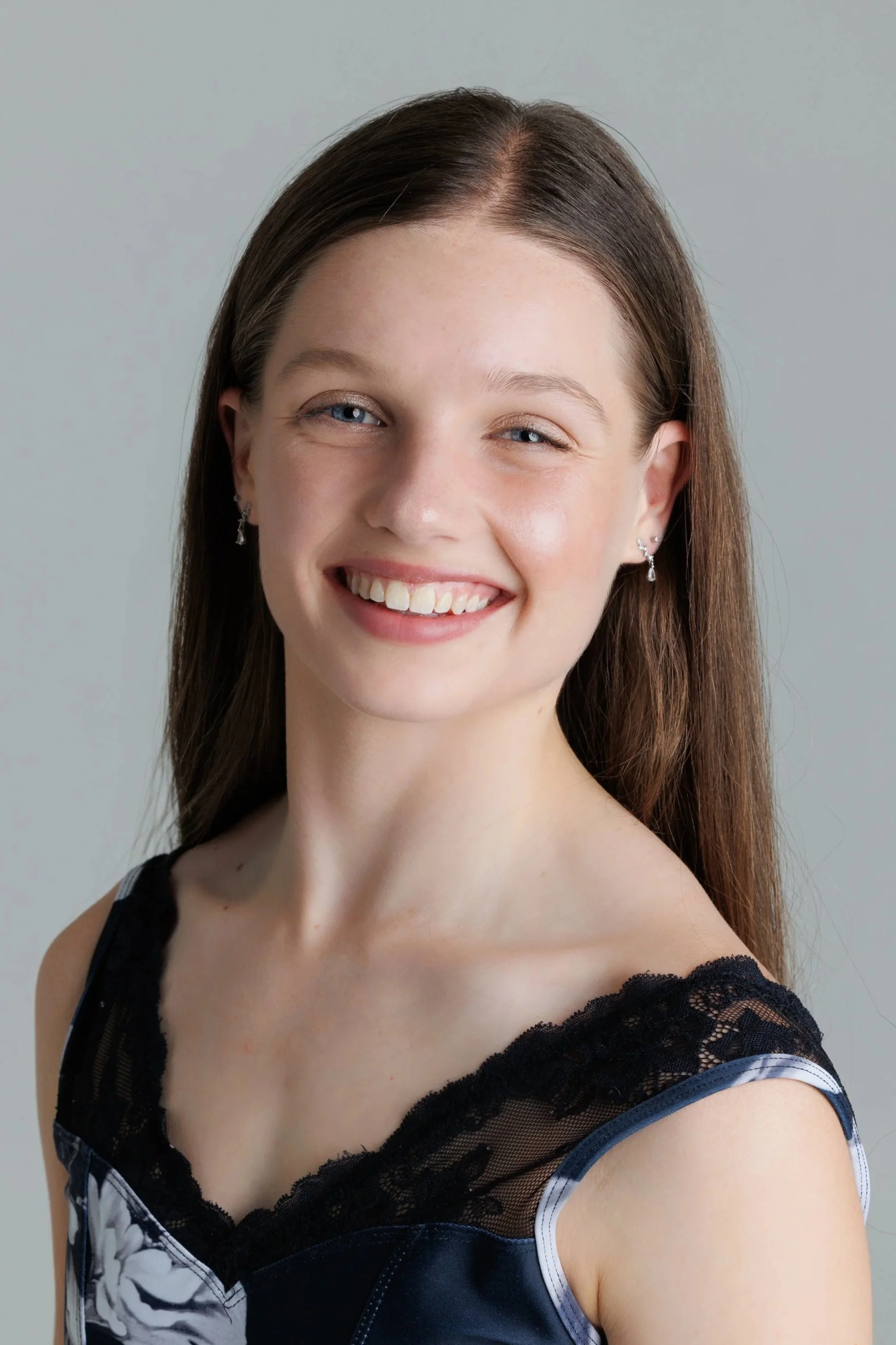 A young woman with long brown hair, blue eyes, and earrings, smiling at the camera. She is wearing a black and white floral dress with black lace trim on the shoulders.