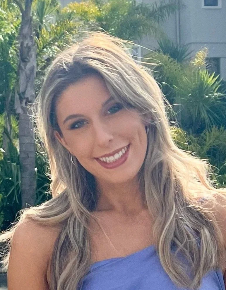 A smiling woman who is a dance teacher at Arc Dance school in Grey Lynn, Auckland, with long wavy blonde hair posing outdoors with green palm trees and plants in the background.