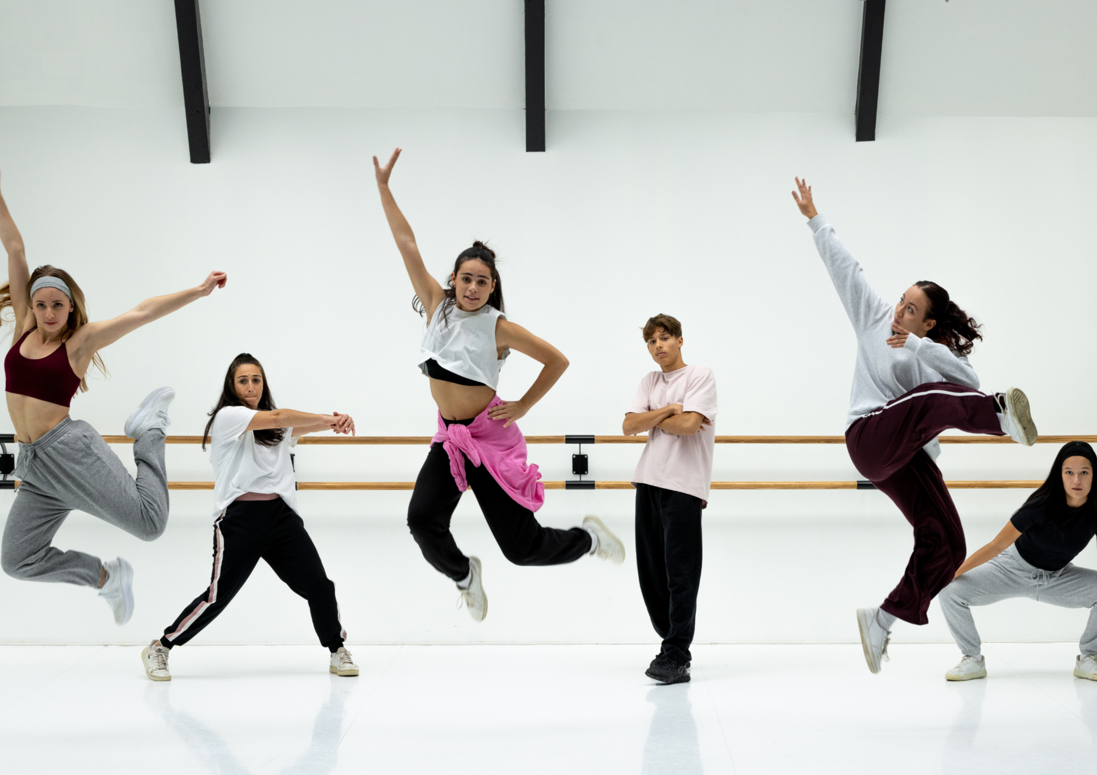Six young dancers practicing in a dance studio with a white wall and a ballet barre, some jumping with arms and legs extended, others standing or crouching, wearing casual dancewear.