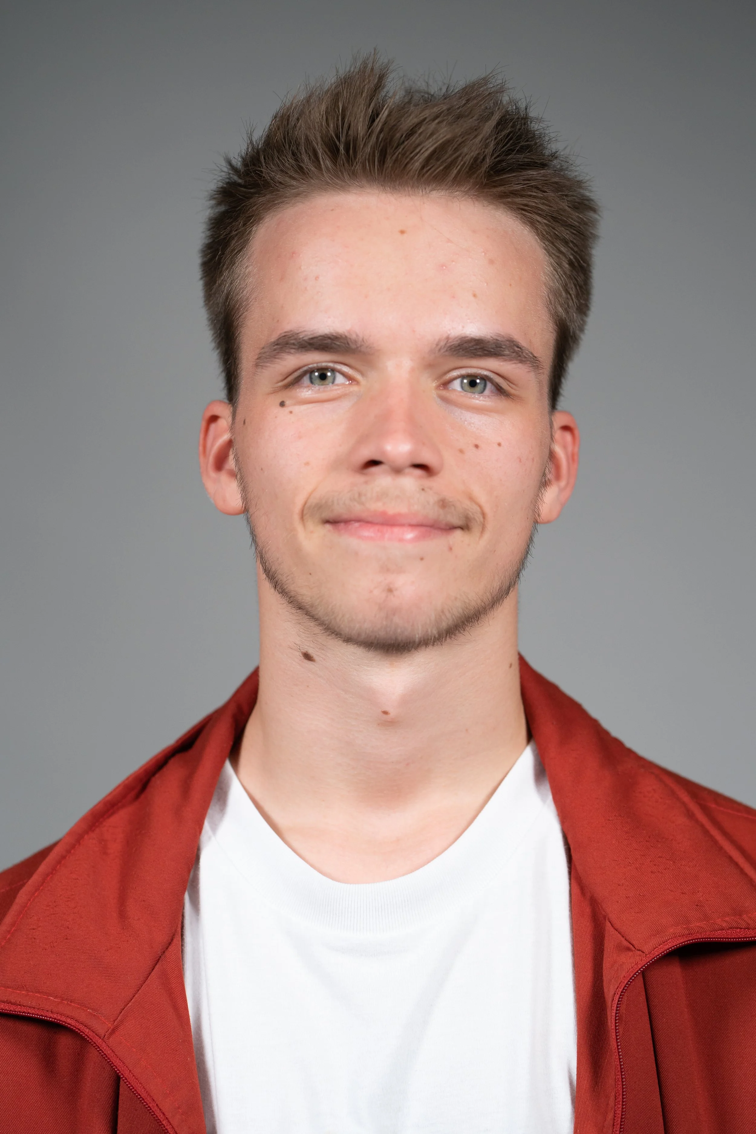 Portrait of a young man who is a dance teacher at Arc Dance school in Grey Lynn, Auckland, with light skin, short light brown hair, and blue eyes, smiling slightly, wearing a white t-shirt and a reddish-brown jacket against a gray background.