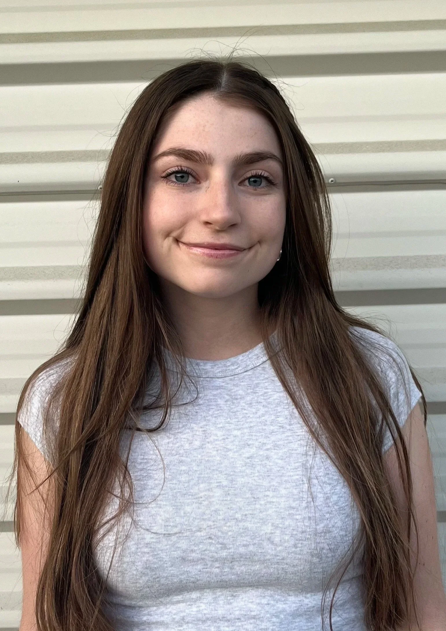A young woman who is a dance teacher at Arc Dance school in Grey Lynn, Auckland, with long brown hair, fair skin, and blue eyes, smiling, wearing a light gray T-shirt standing in front of a white wall with horizontal siding.