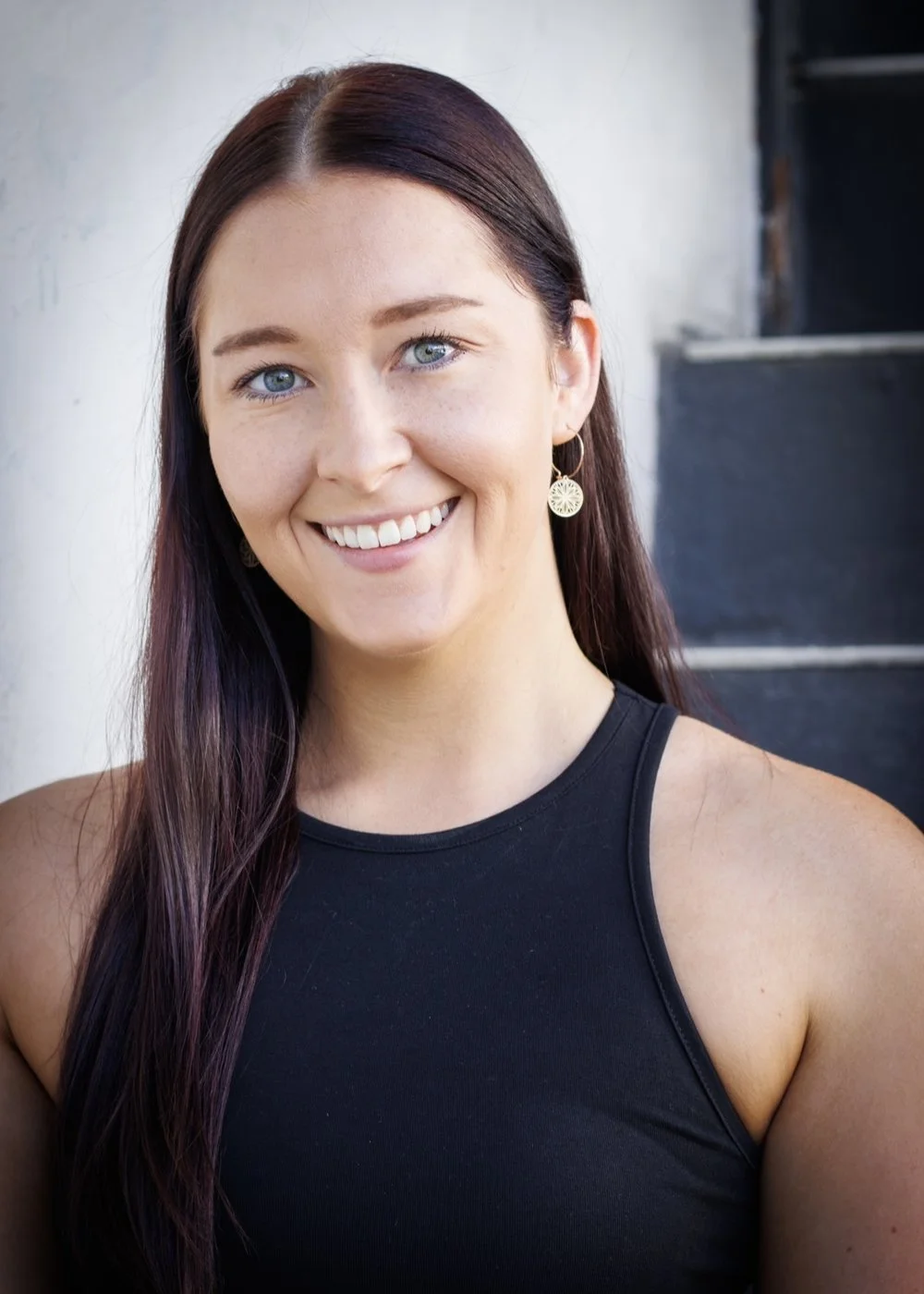 Close-up of a young woman who is a dance teacher in Grey Lynn, Auckland, with long dark hair, blue eyes, wearing a black sleeveless top and gold earrings, smiling outdoors against a blurred background.
