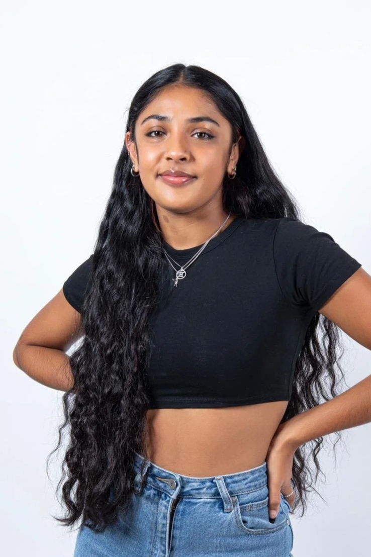 A young woman who is a dance teacher at Arc Dance school in Grey Lynn, Auckland, with long, wavy black hair wearing a black crop top, blue jeans, silver jewelry, and standing against a plain white background.
