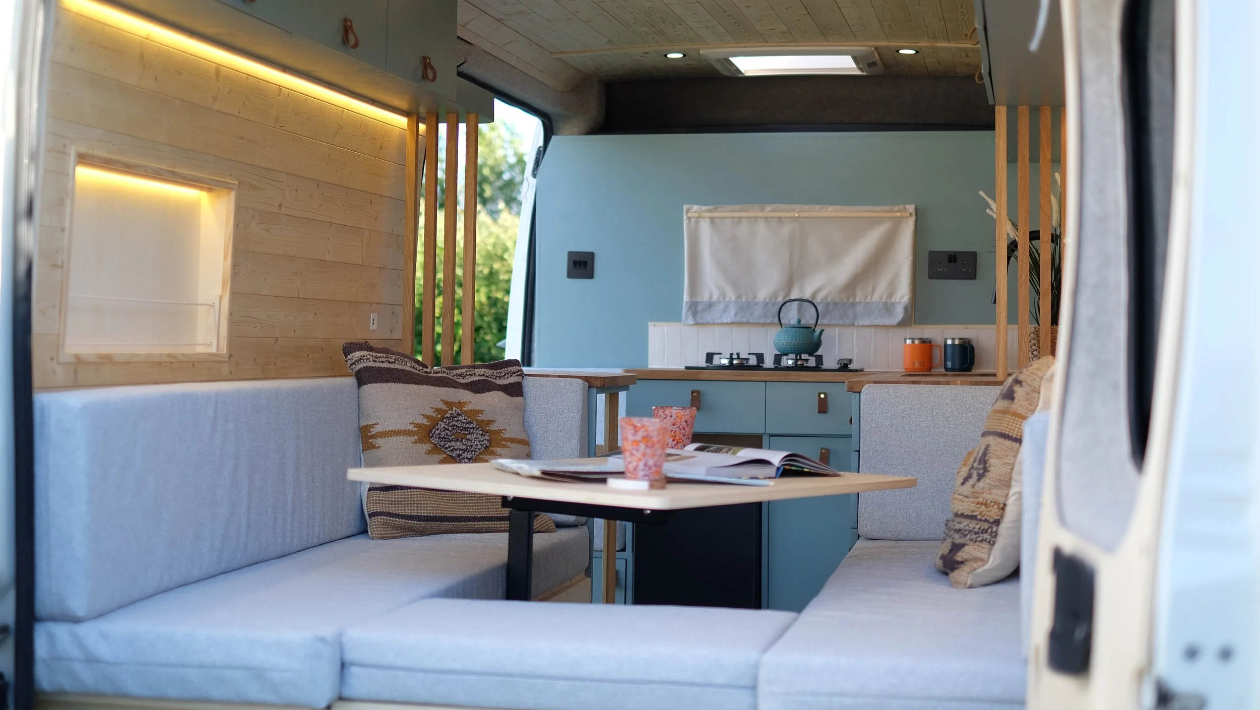 Interior of a camper van showing a cozy living area with a round table, built-in sofa with decorative pillows, and a compact kitchenette with a blue counter, kettle, mugs, and a window with a curtain.