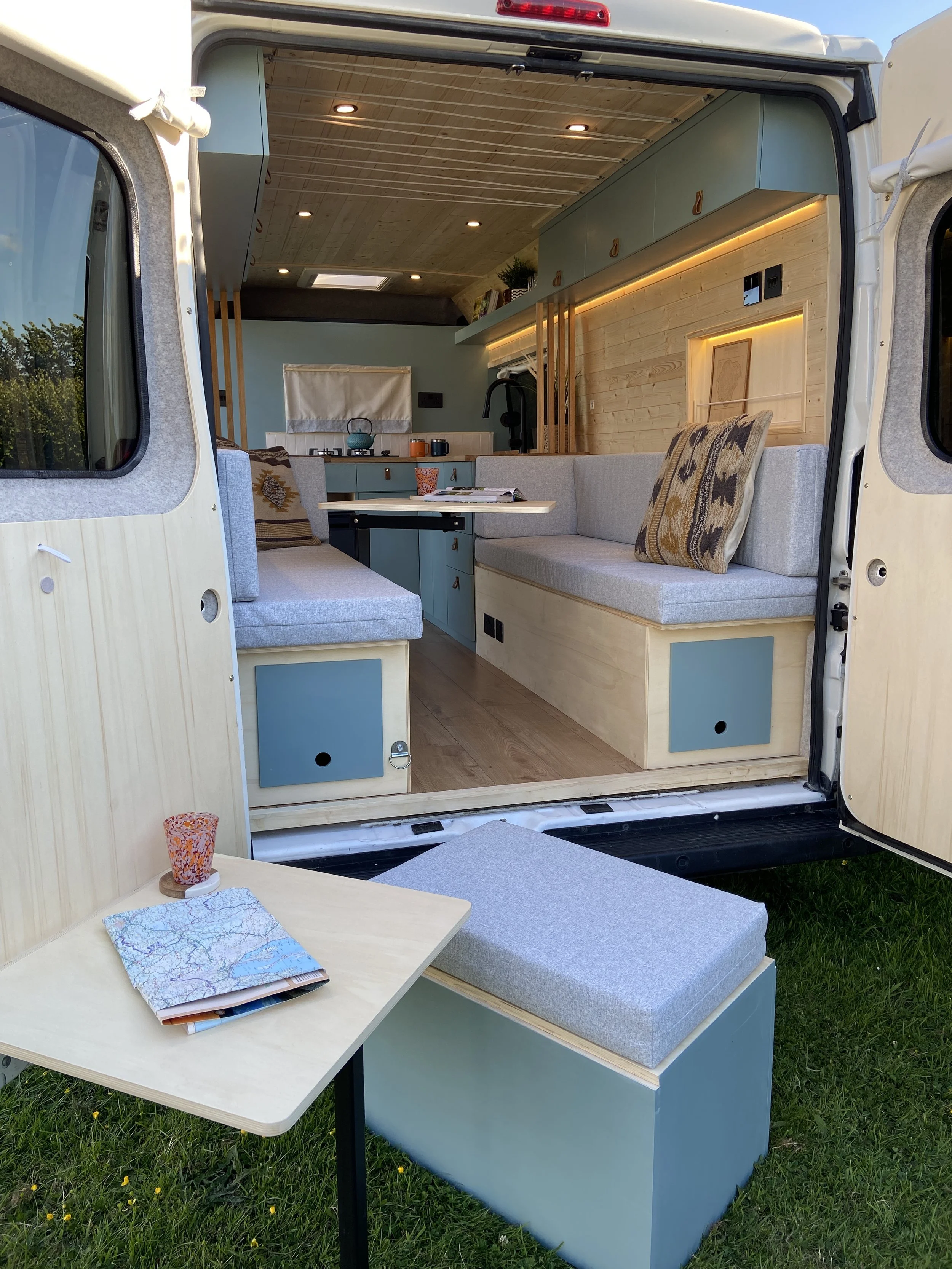Interior of a compact camper van with a built-in seating area, kitchen, and wooden ceiling and walls, set outdoors on green grass.