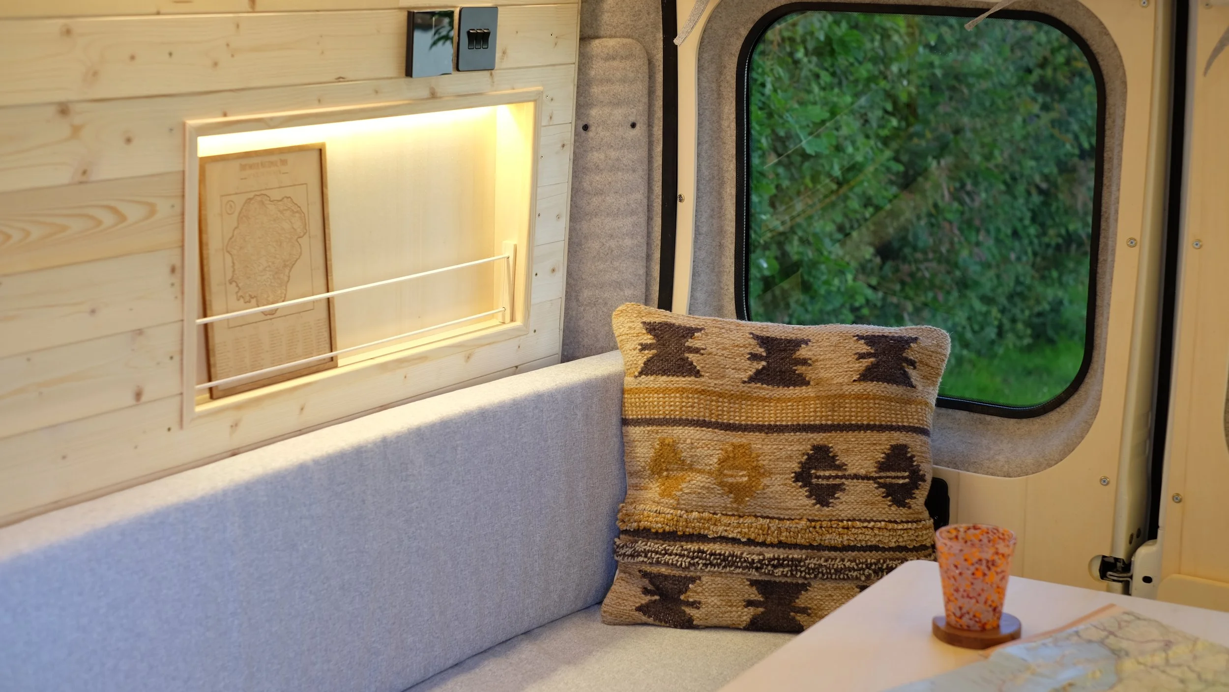 The image shows the interior of a camper van with a light gray cushioned seating area, a decorative throw pillow with a tribal pattern, a window with a view of green trees, a small table with a speckled drink in a cup, and a framed map on a wooden wa