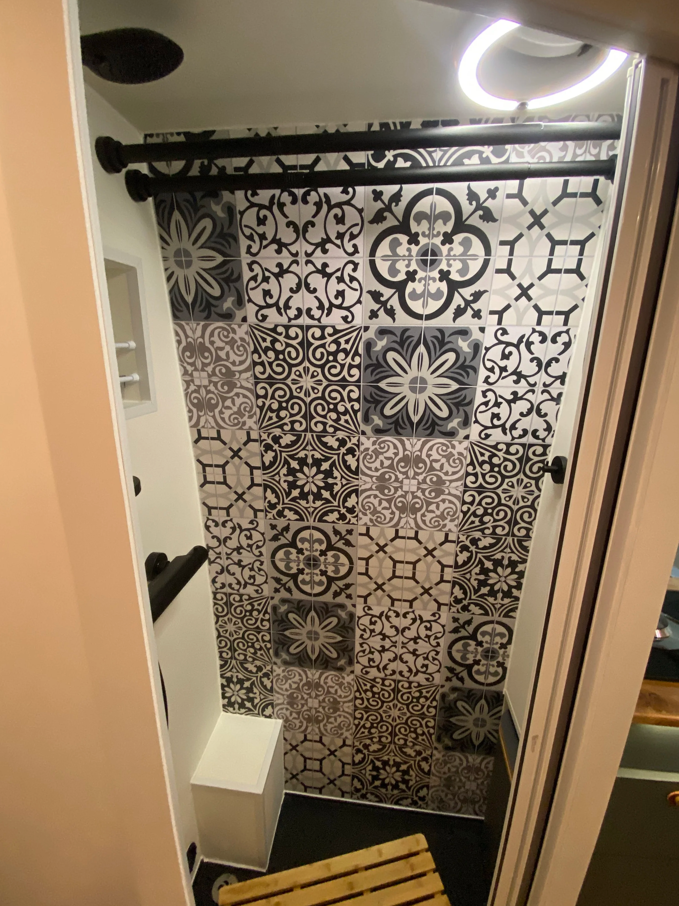 Small shower with patterned black and white decorative tiles, black grab bar, two black curtain rods, small built-in niche, circular ceiling light, and a wooden slatted bench.