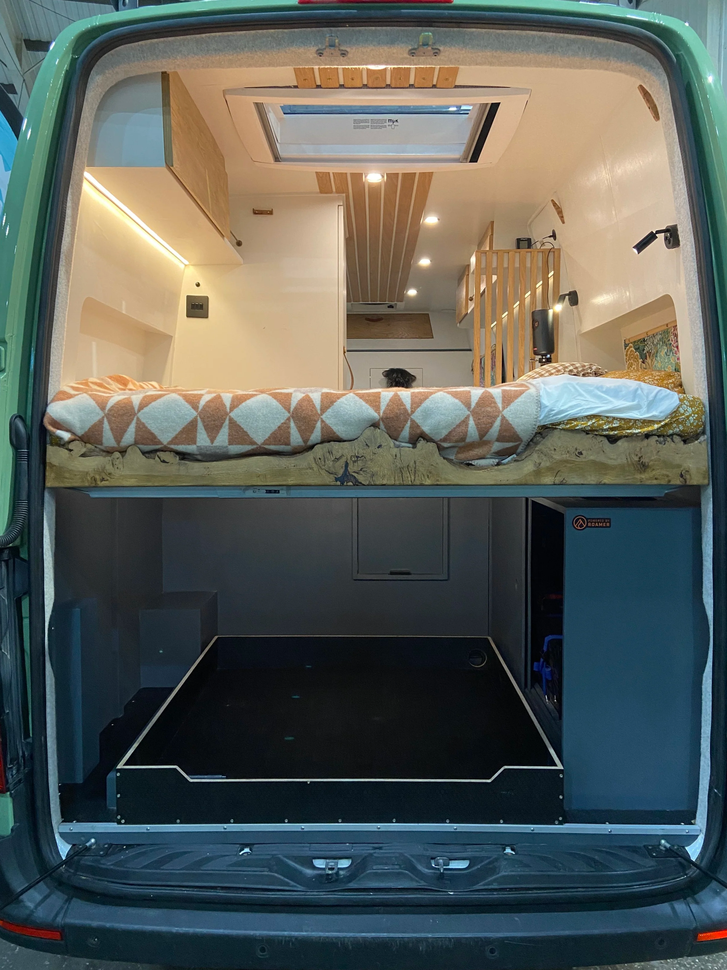 Interior of a converted camper van with a bed and storage area underneath. The bed is made with a geometric-patterned blanket, and there is a view of the ceiling and walls with wooden accents and lighting.