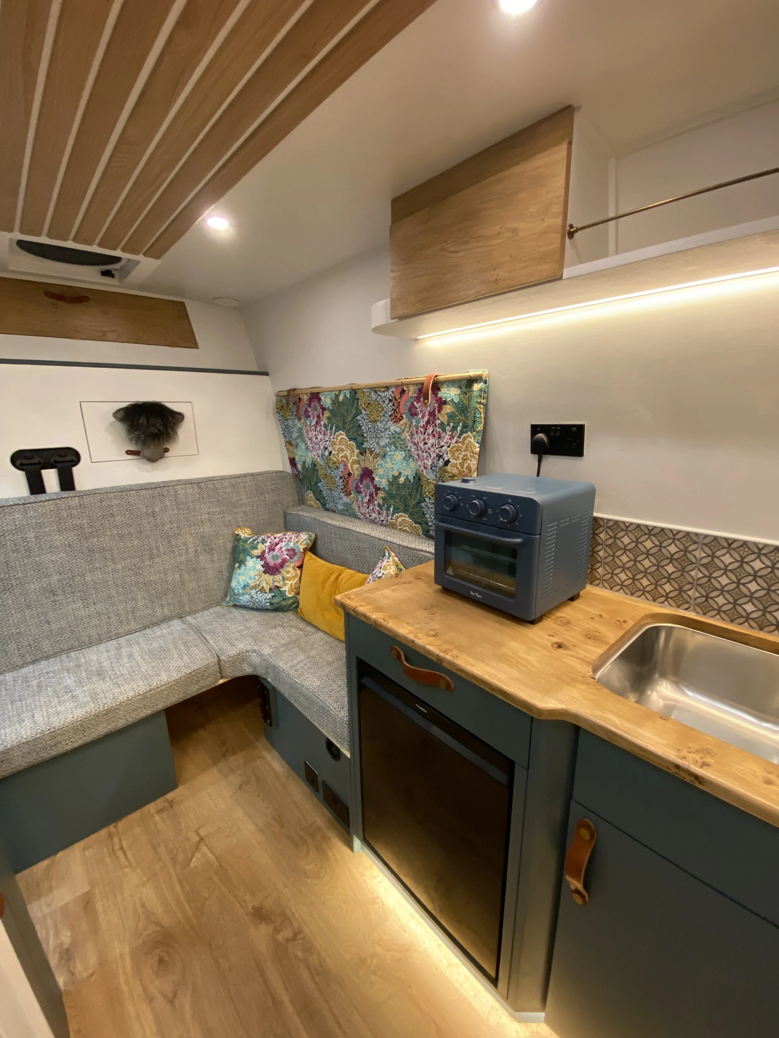 Tiny kitchen with a curved gray couch against the wall, colorful floral pillow, small wooden countertop with a mini oven, sink, and kitchenette appliances, decorative patterned backsplash, floral curtain, wooden storage cabinets, and ceiling with rec