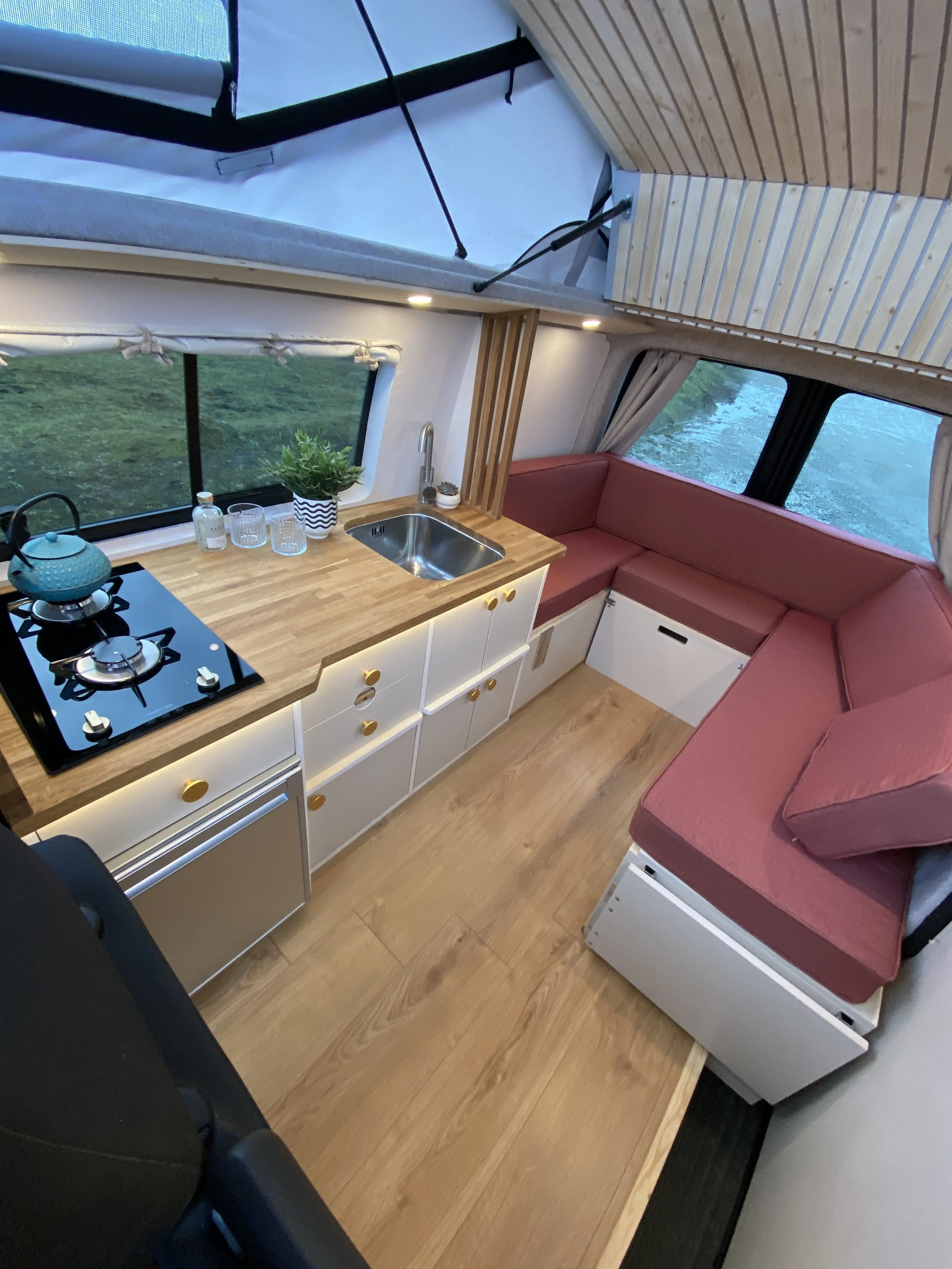 Interior of a compact camper van with a small kitchen area and V-shaped seating with pink cushions, window view of greenery, wooden accents, and a ceiling with wood paneling.