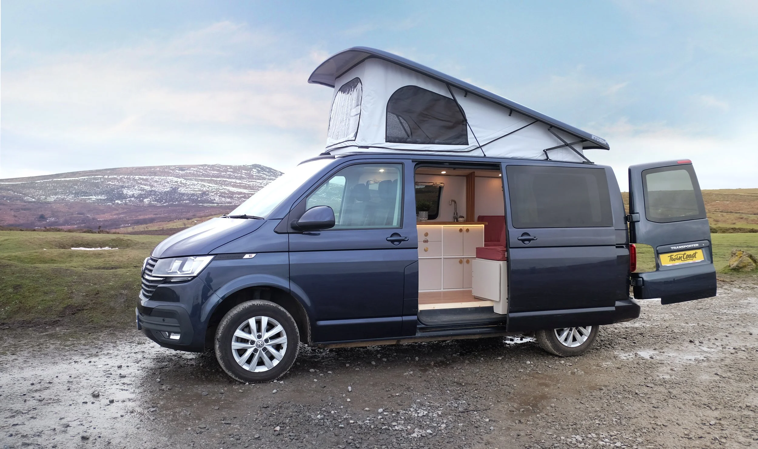 Black campervan conversion with a pop-up roof, parked outdoors with a mountain and grassy landscape in the background. The side door is open, revealing the interior kitchen and seating area.