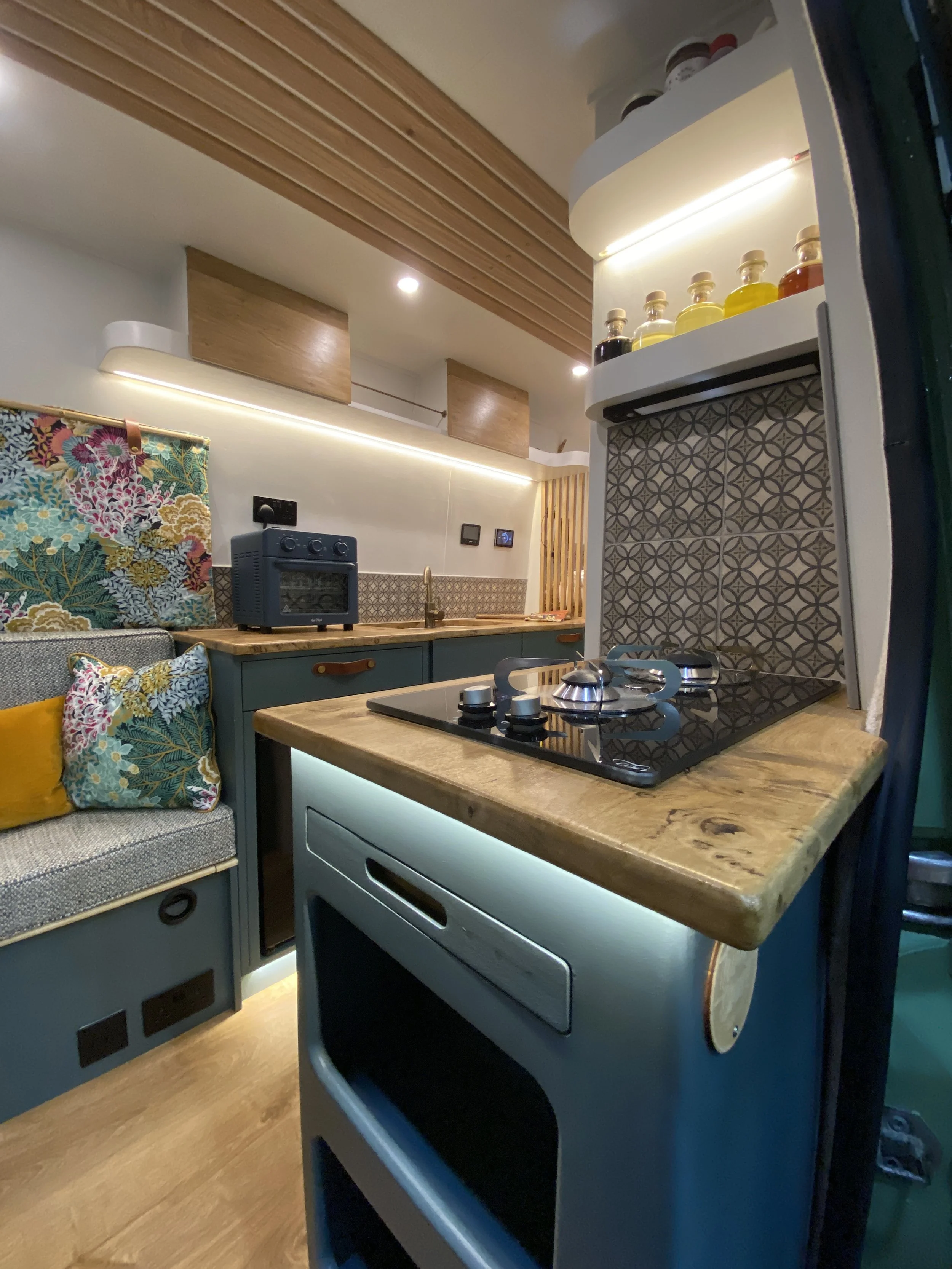 Van conversion kitchen corner with a wooden countertop, a stovetop, patterned tile backsplash, colourful cushions on a cozy seating area, and shelves with bottles, all in a modern, stylish design.