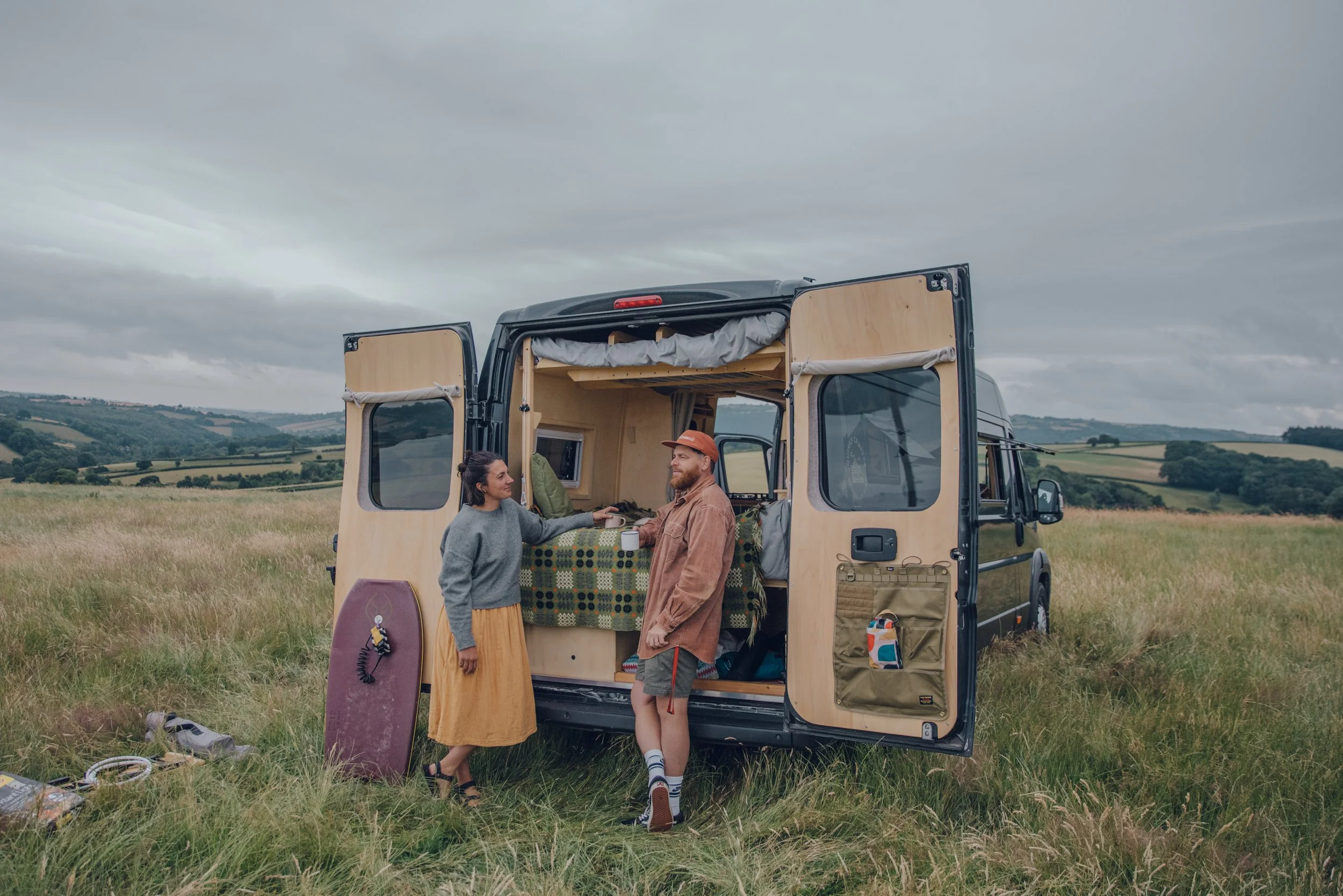 Two people standing outside a camper van in a grassy field, with overcast sky, engaging in conversation with each other.